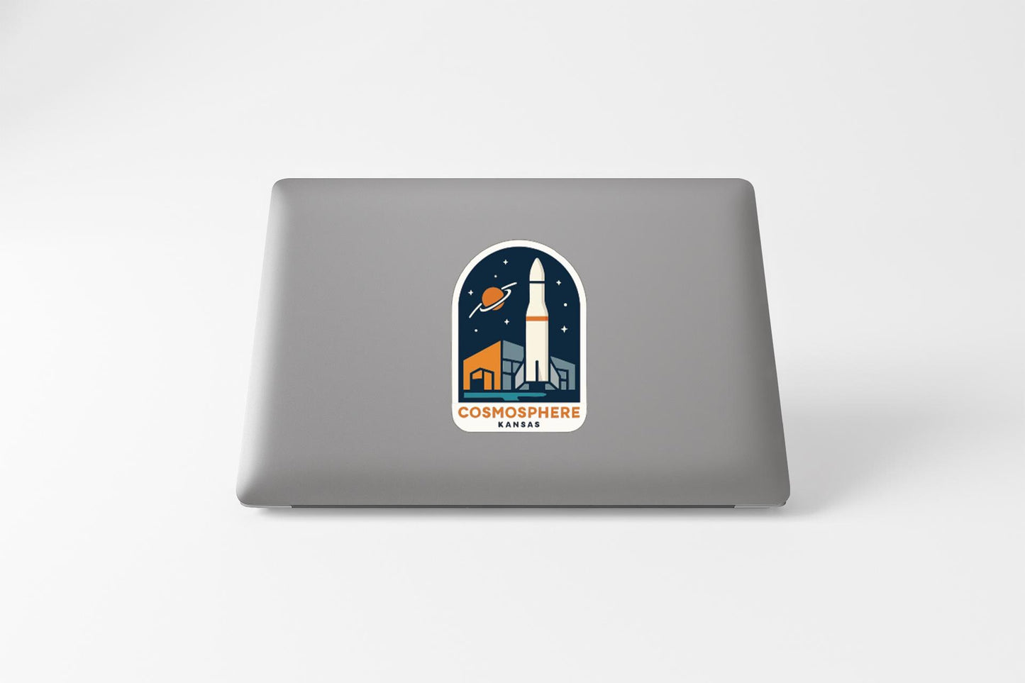 a sticker on the back of a laptop that displays a rocket ship and a planet, along with the text "COSMOSPHERE KARA".