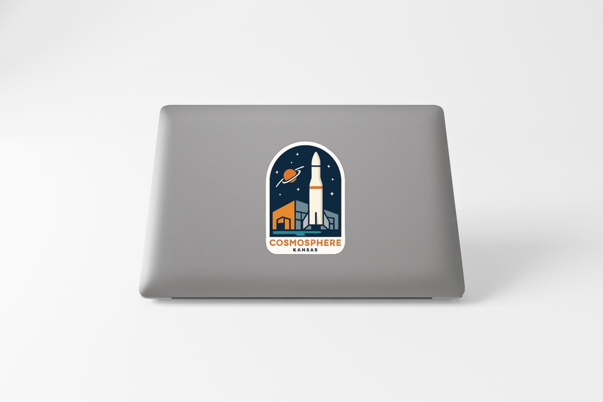 a sticker on the back of a laptop that displays a rocket ship and a planet, along with the text "COSMOSPHERE KARA".