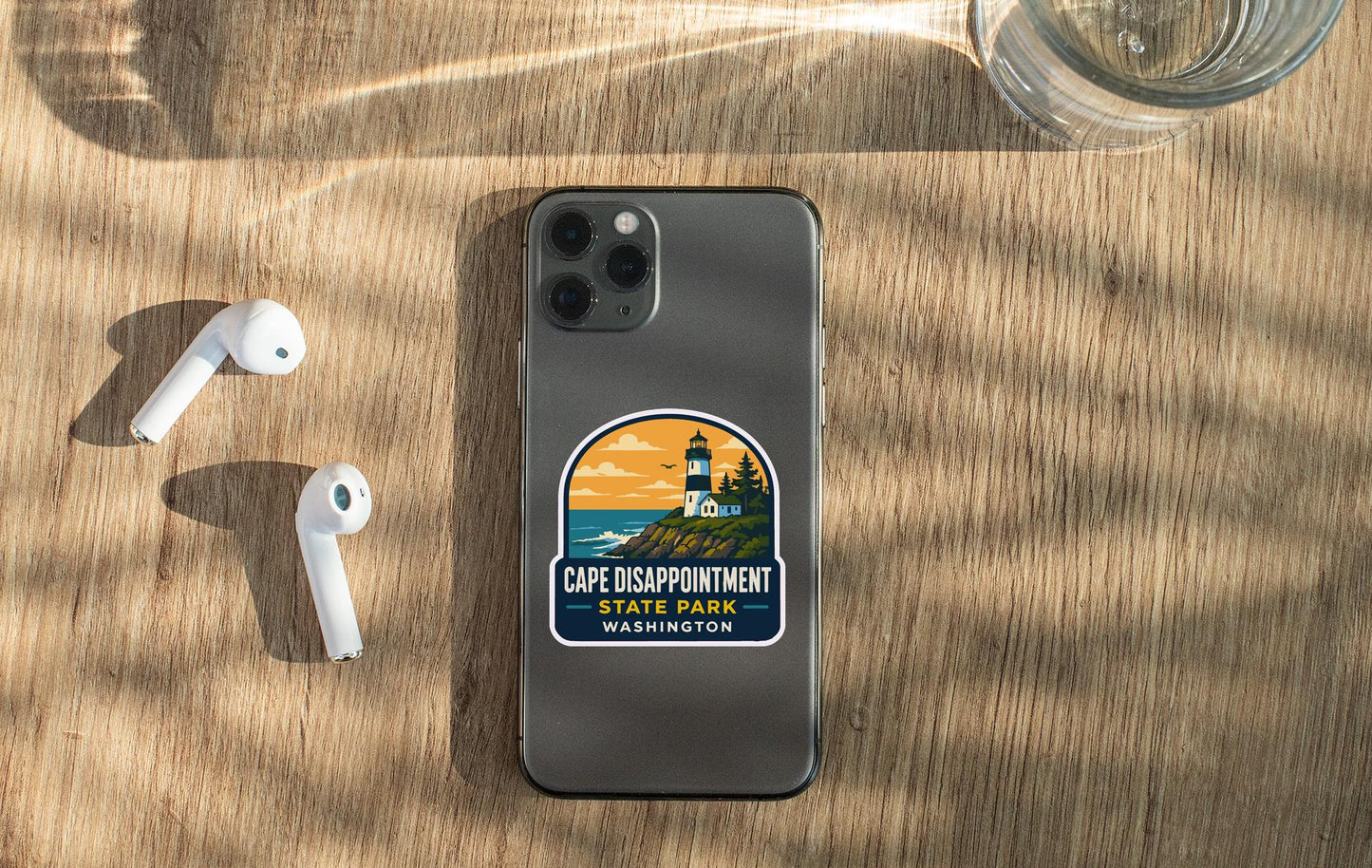 a smartphone with a sticker on it, placed on a wooden surface alongside a pair of white earbuds and a glass of water.