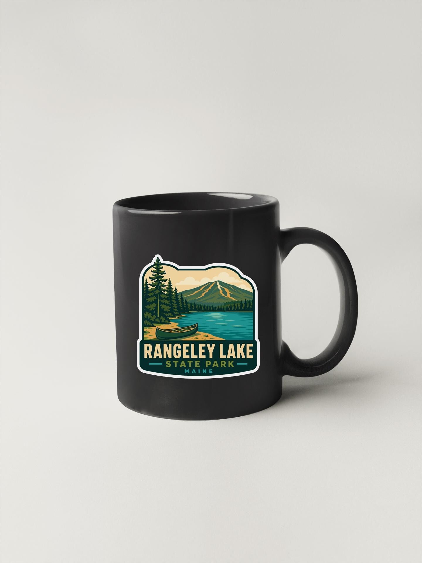 A black coffee mug with a graphic design depicting a scenic landscape featuring a lake, mountains, and pine trees.