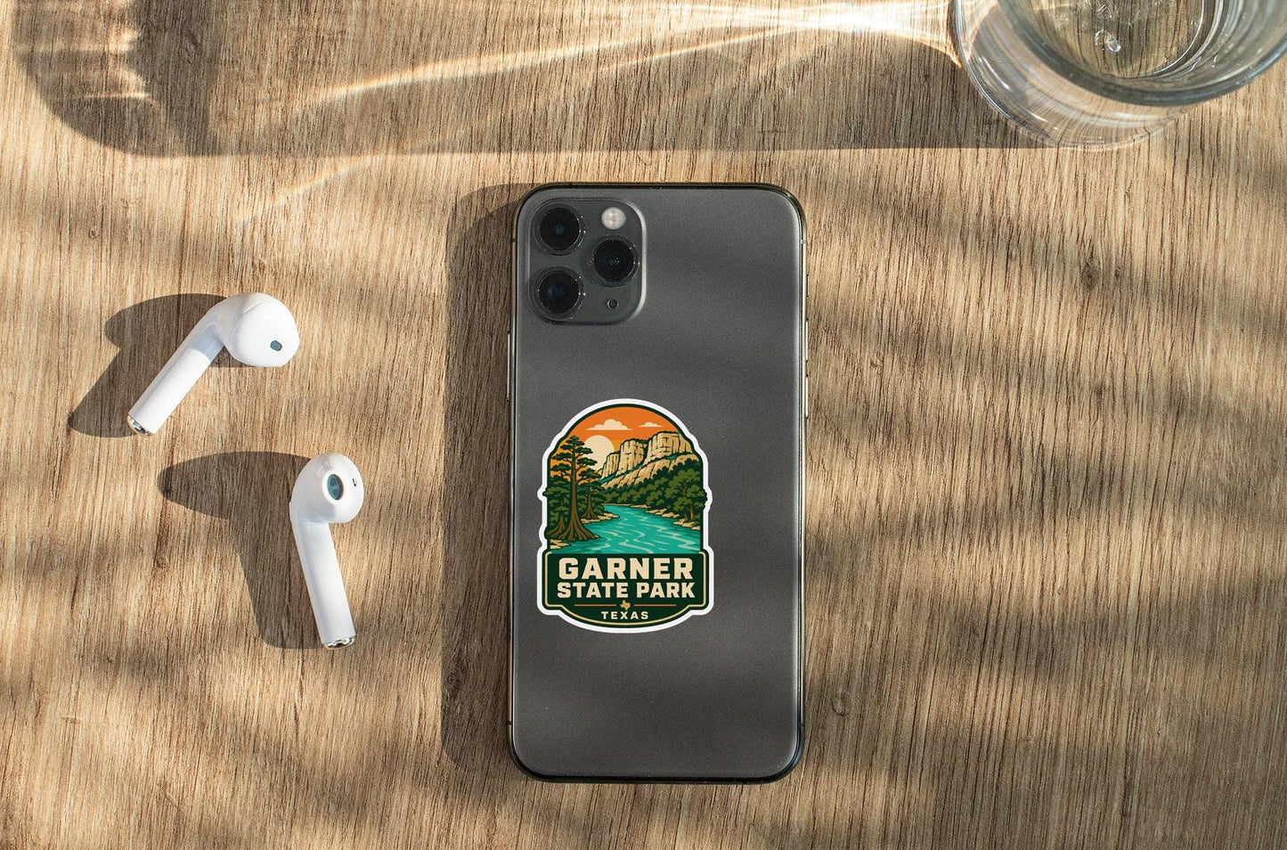 a smartphone with a sticker on it, placed on a wooden surface alongside a pair of white earbuds and a glass of water.
