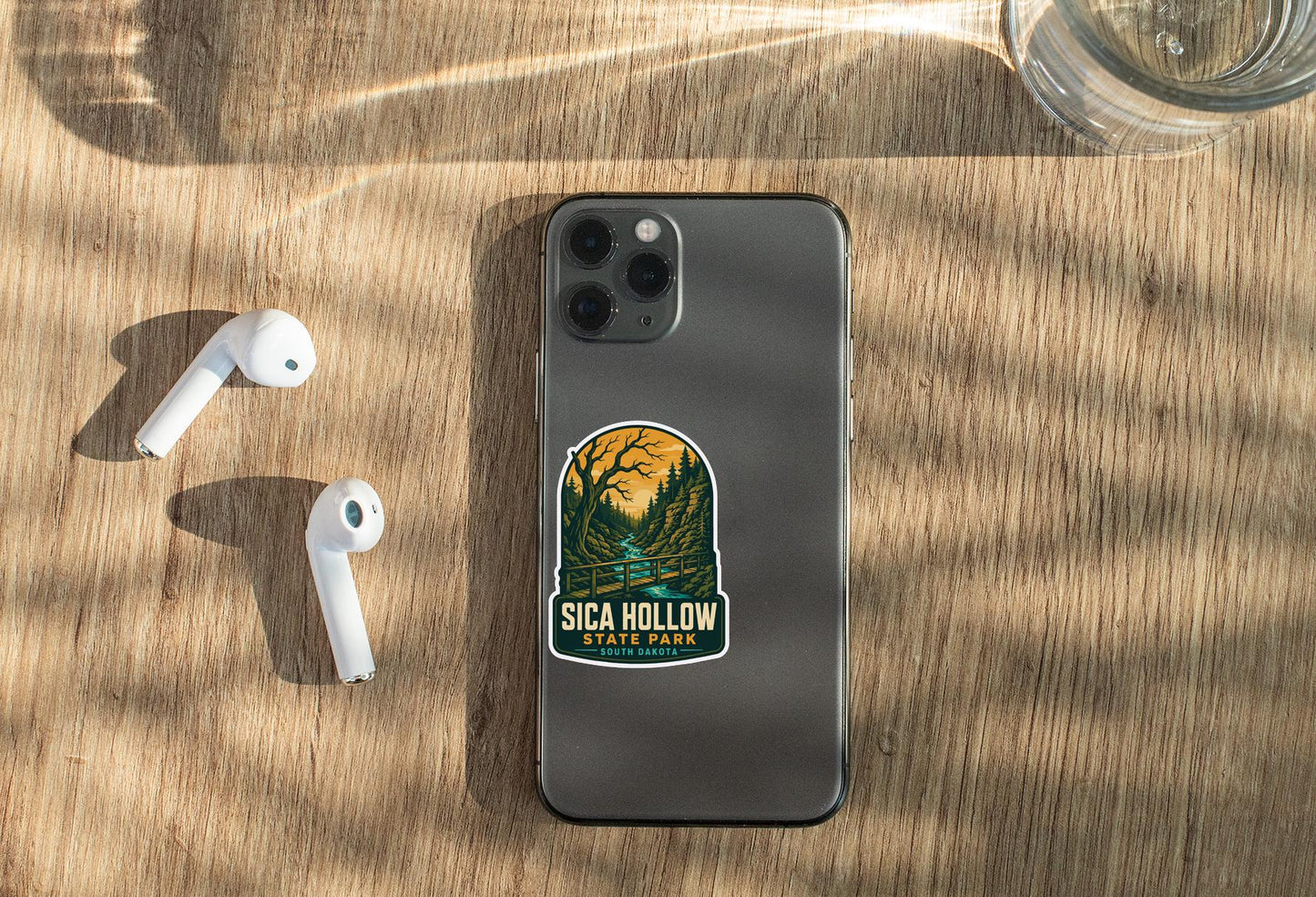 a smartphone with a sticker on it, placed on a wooden surface alongside a pair of white earbuds and a glass of water.