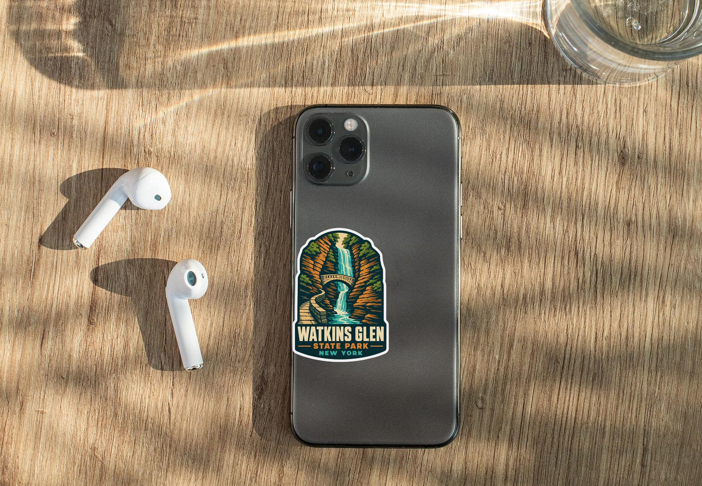 a smartphone with a sticker on it, placed on a wooden surface alongside a pair of white earbuds and a glass of water.