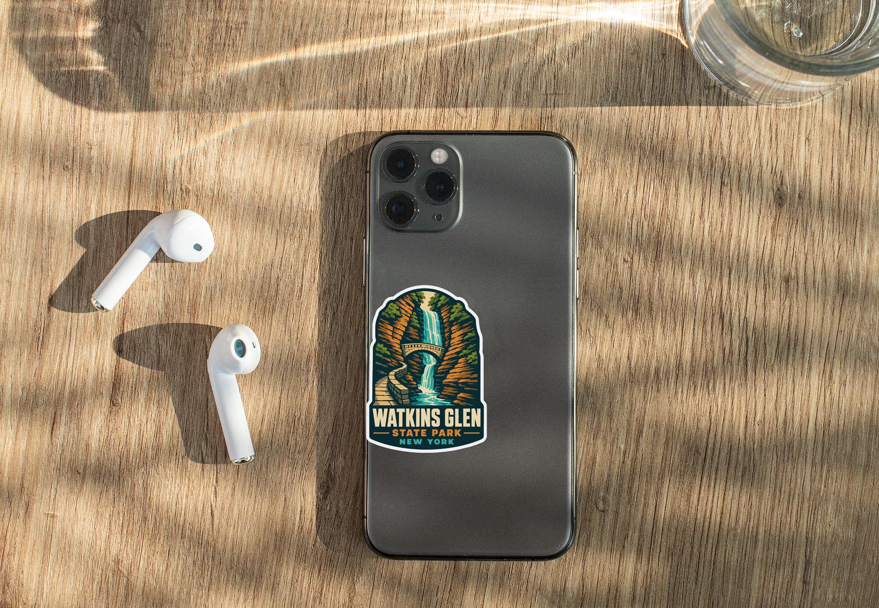 a smartphone with a sticker on it, placed on a wooden surface alongside a pair of white earbuds and a glass of water.