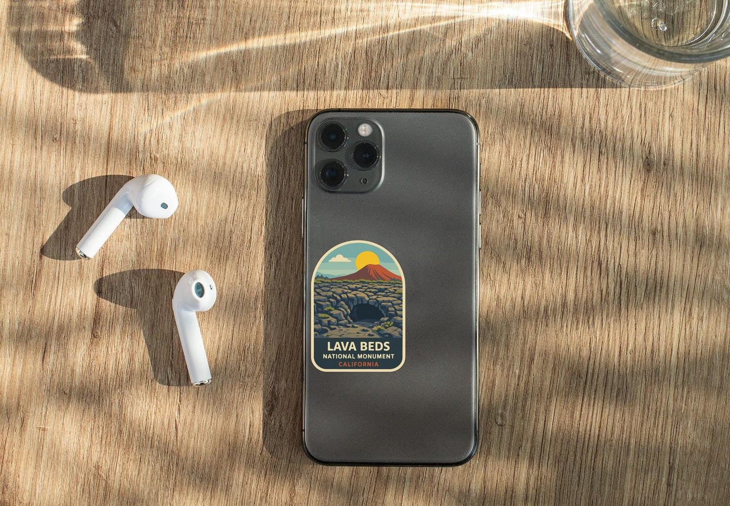 a smartphone with a sticker on the back, placed on a wooden surface alongside a pair of white earbuds and a glass of water.