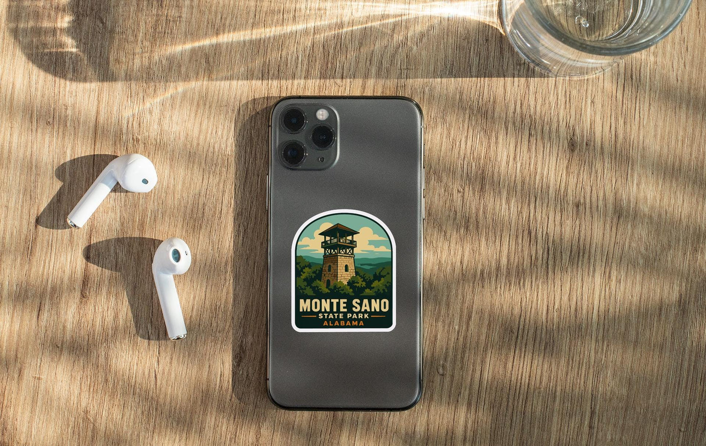 a smartphone with a sticker on the back, placed on a wooden surface alongside a pair of white earbuds and a glass of water.