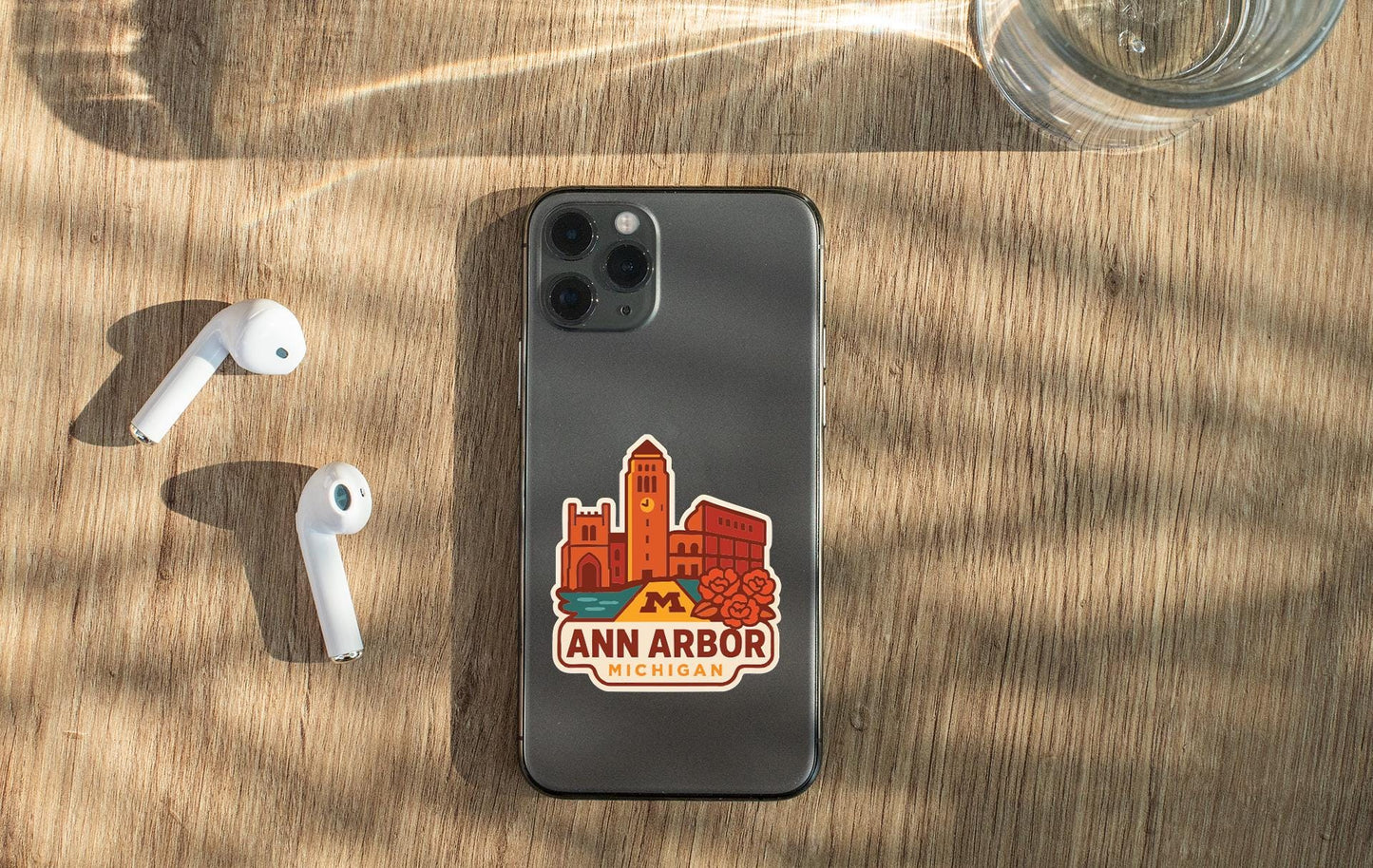 a smartphone with a sticker of Ann Arbor, Michigan, placed on a wooden surface alongside a pair of white earbuds and a glass of water.