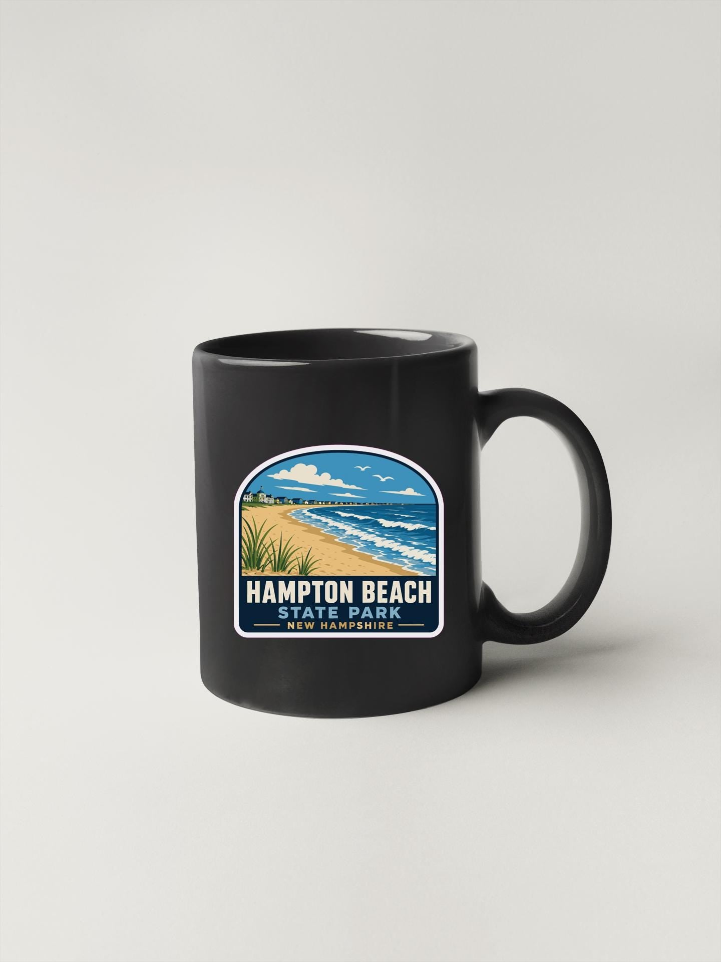 A black coffee mug with a graphic design depicting a beach scene and the text "HAMPTON BEACH STATE PARK NEW HAMPSHIRE".