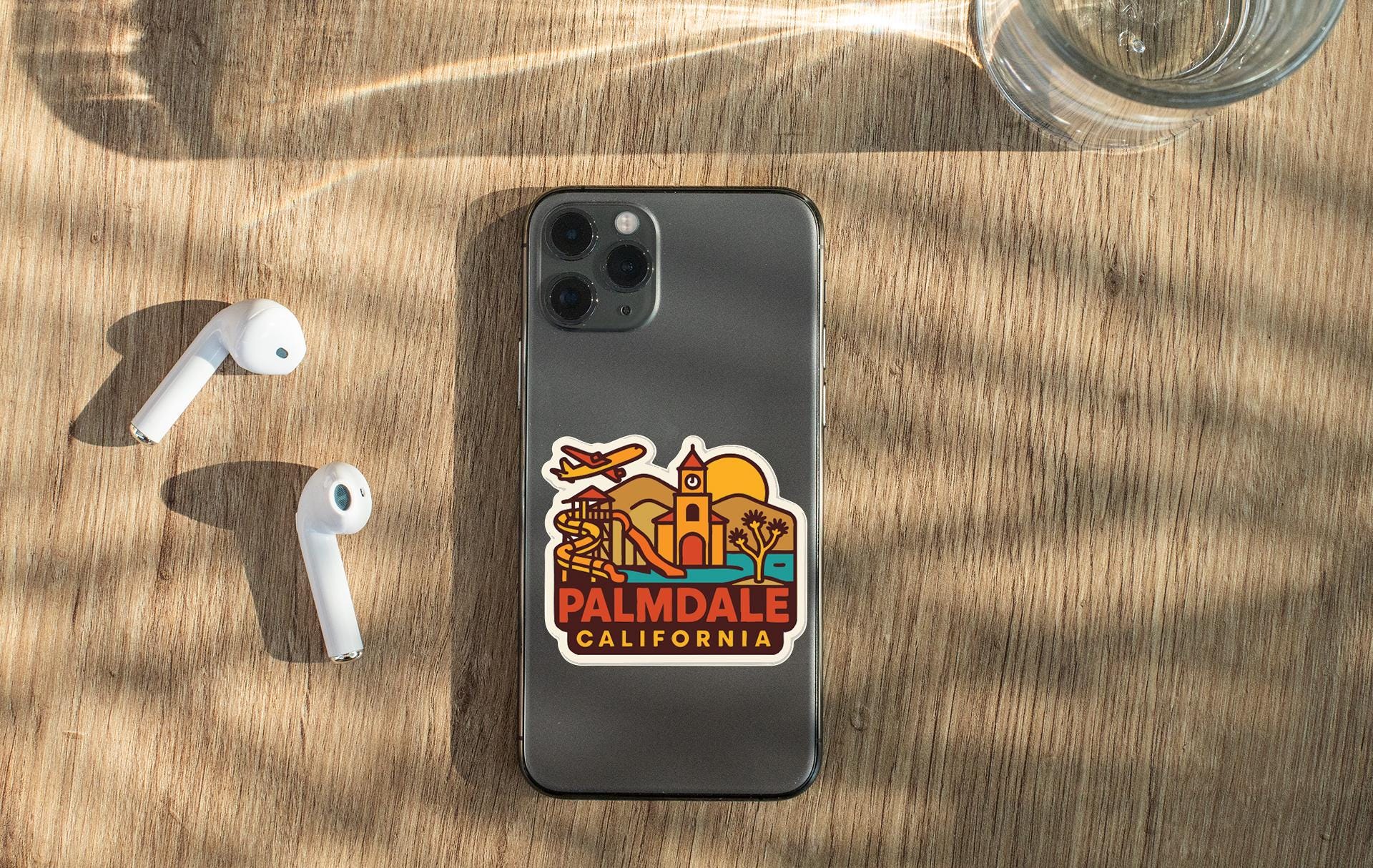 a smartphone with a sticker on the back, placed on a wooden surface alongside a pair of white earbuds and a glass of water.
