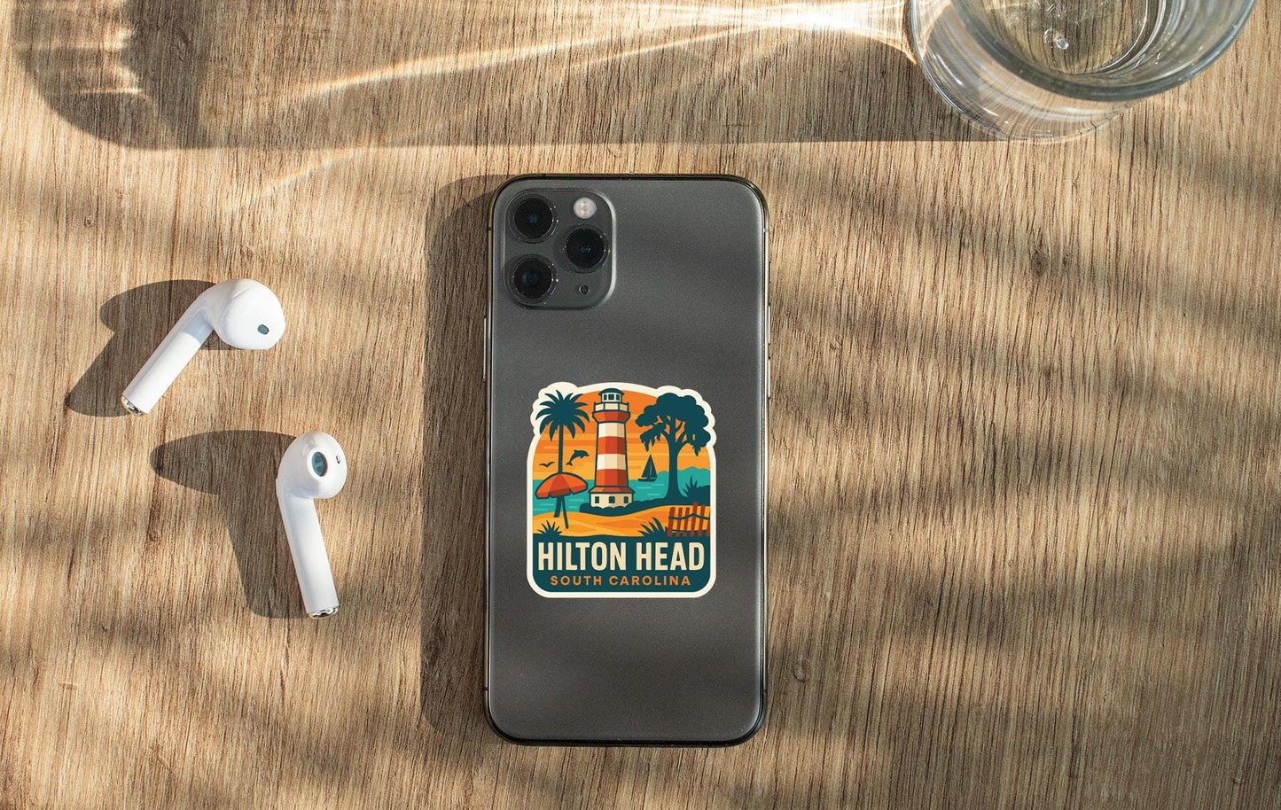 a smartphone with a sticker on the back, placed on a wooden surface alongside a pair of white earbuds and a glass of water.