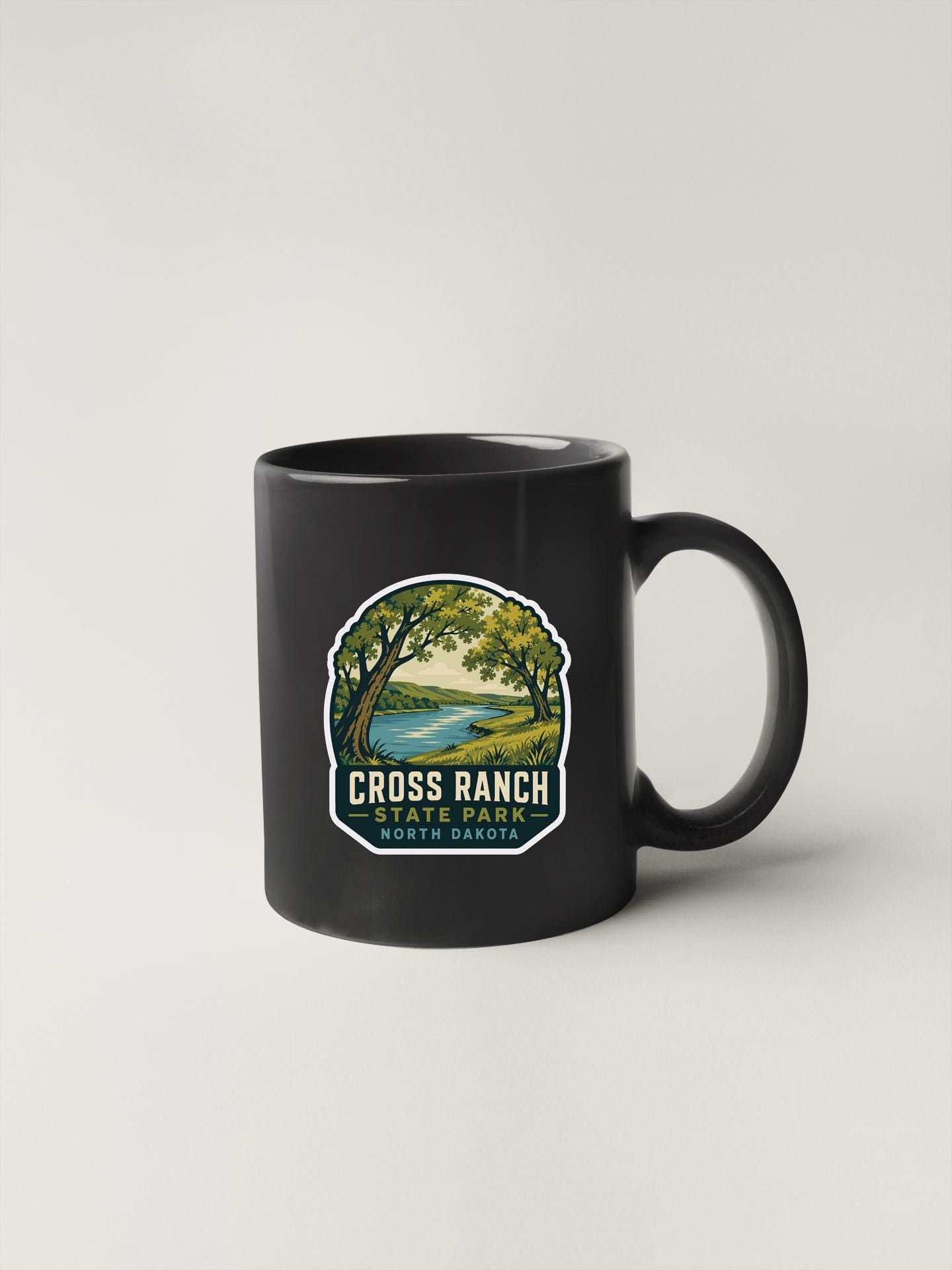 A black coffee mug with a graphic design depicting a scenic landscape, including a river, trees, and a lake.