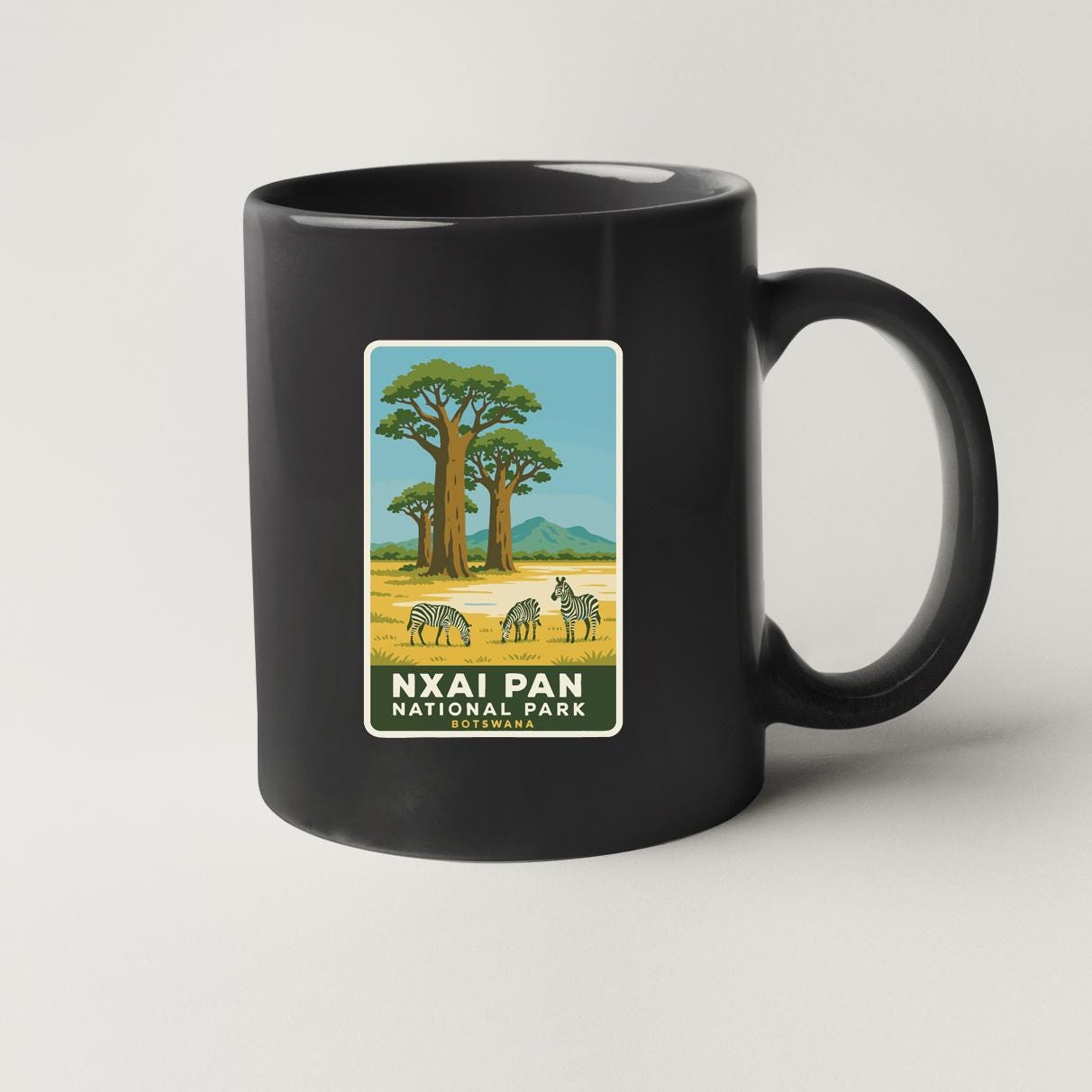 A black coffee mug with a label depicting a scenic landscape with zebras and trees, representing the Nxai Pan National Park.