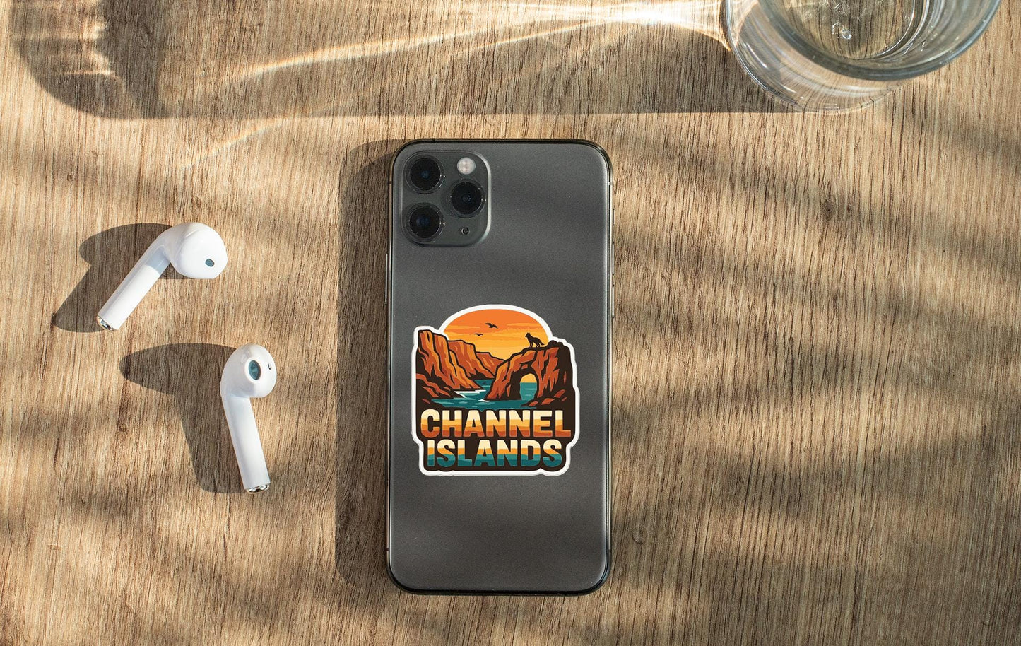 a smartphone with a sticker on the back, placed on a wooden surface alongside a pair of white earbuds and a glass of water.