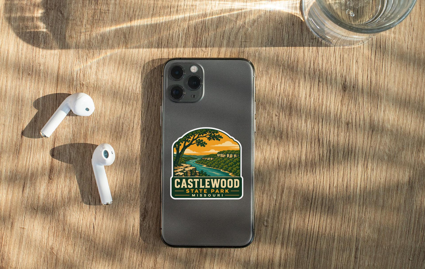 a smartphone with a sticker on it, placed on a wooden surface alongside a pair of white earbuds and a glass of water.
