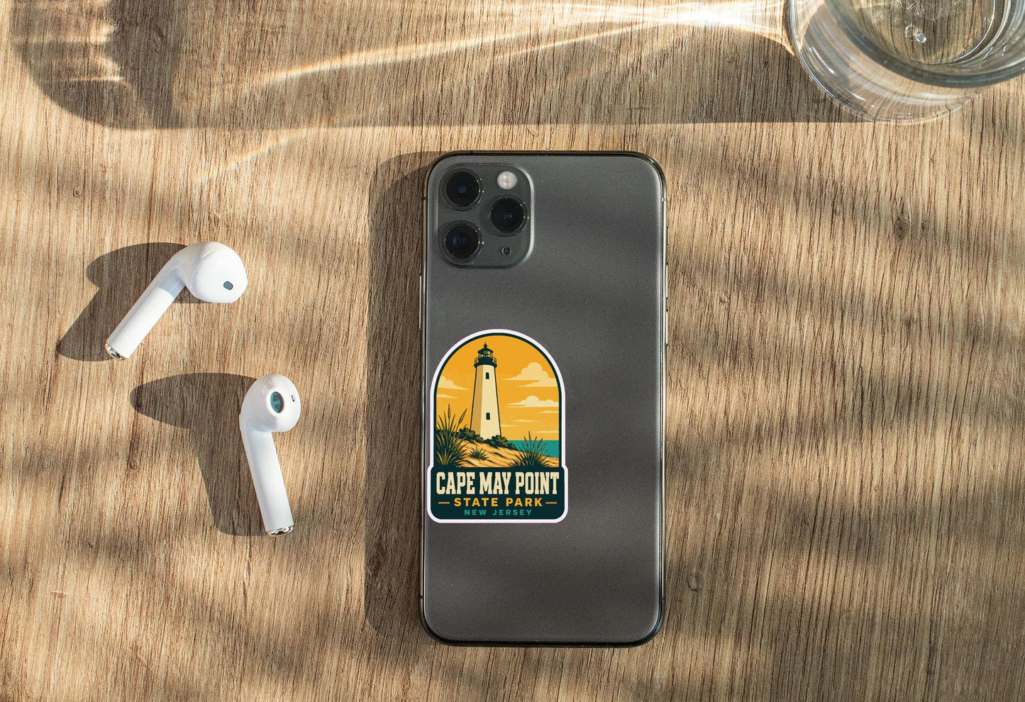 a smartphone with a sticker on the back, placed on a wooden surface alongside a pair of white earbuds and a glass of water.