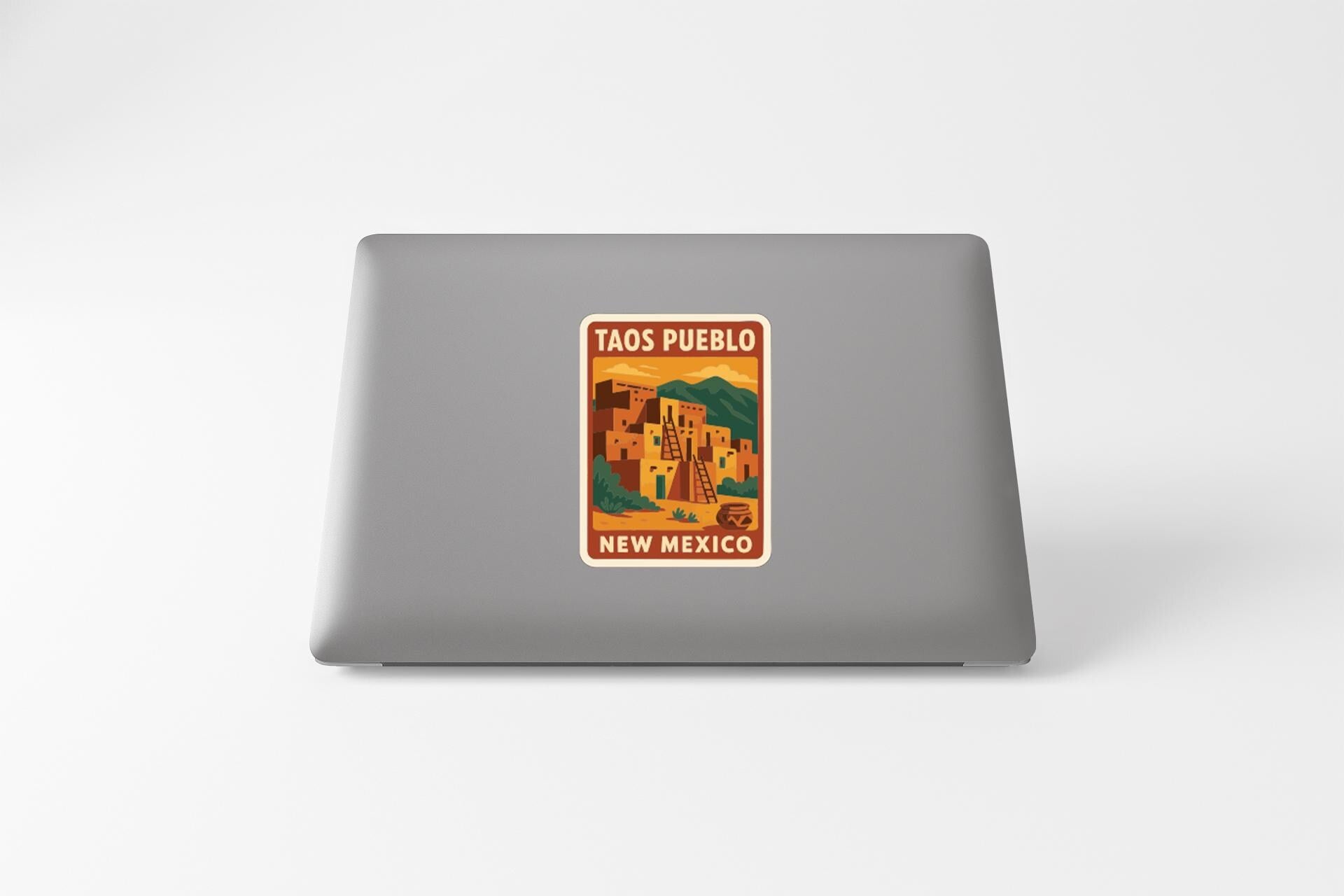 A sticker on the back of a laptop displaying the text "Taos Pueblo, New Mexico" along with an illustration of a desert landscape.