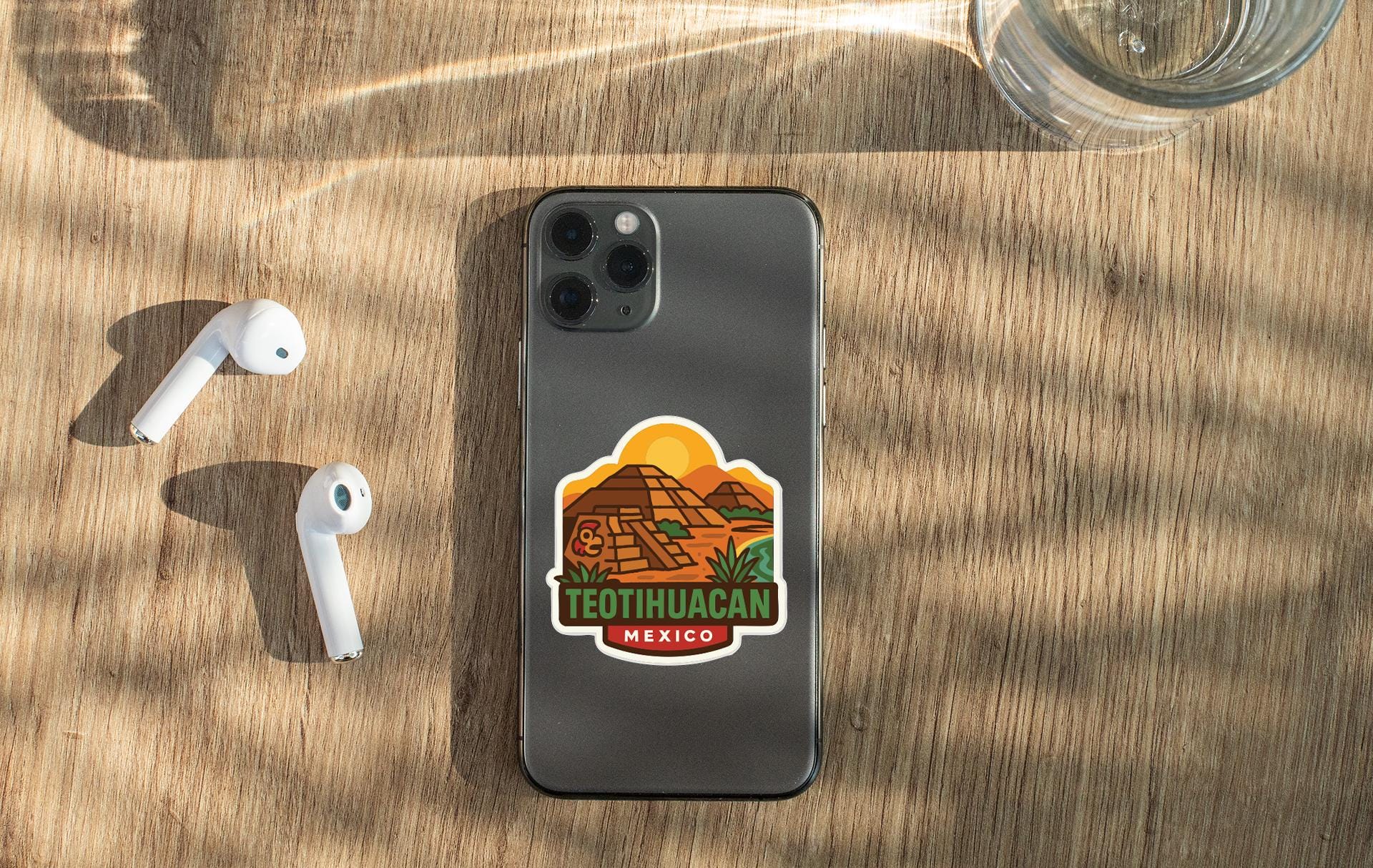 a smartphone with a sticker on the back, placed on a wooden surface alongside a pair of white earbuds and a glass of water.