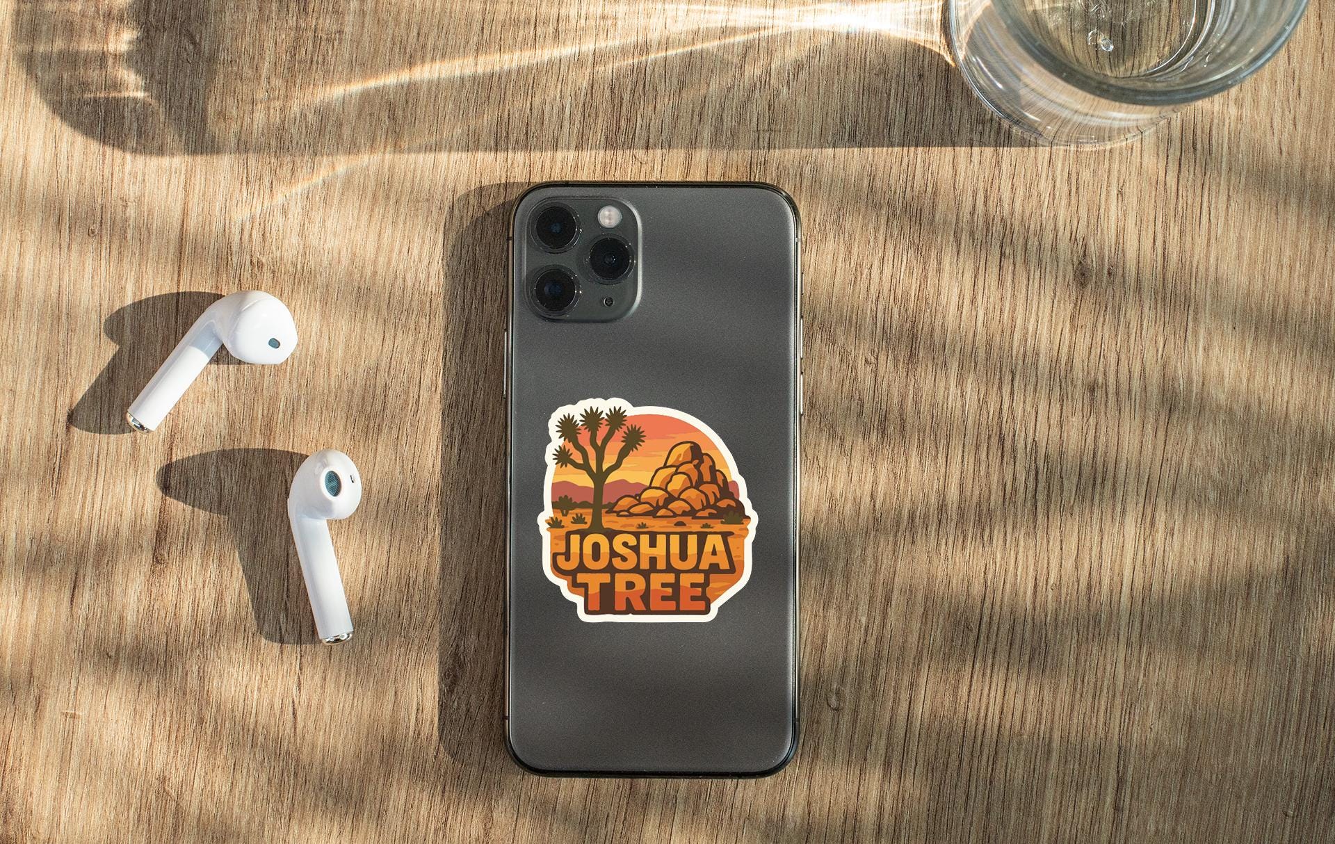 a smartphone with a sticker on the back, along with a pair of white earbuds and a glass of water on a wooden surface.