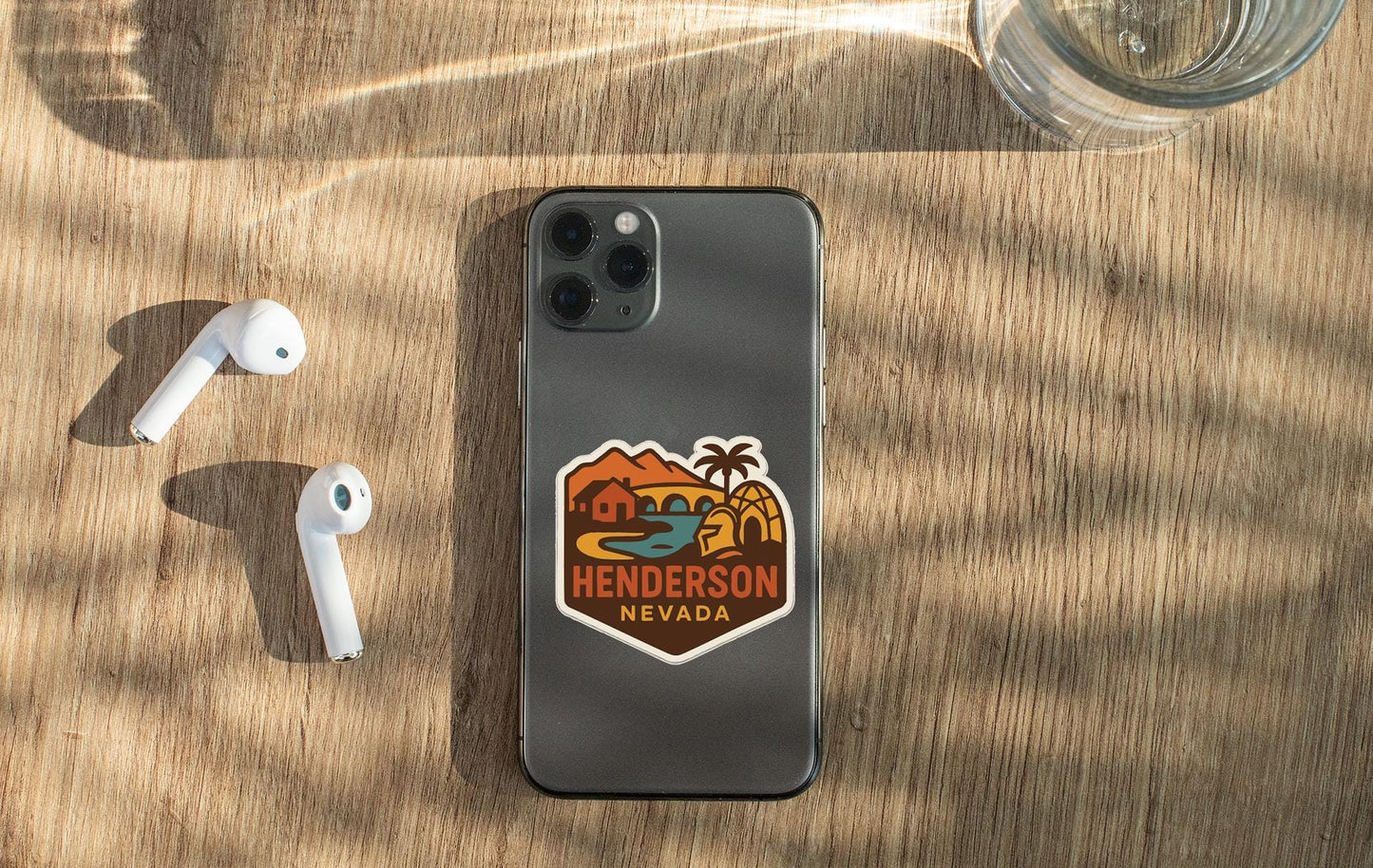 a smartphone with a sticker on the back, a pair of white earbuds, and a glass of water on a wooden surface.