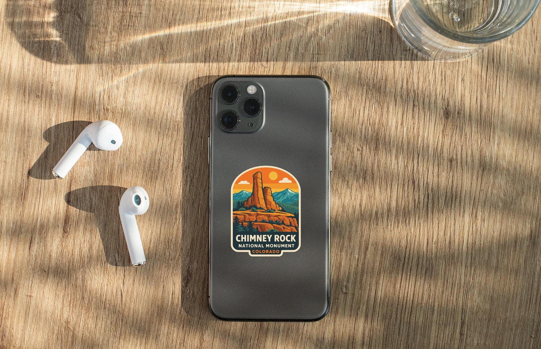 a smartphone with a sticker on the back, placed on a wooden surface alongside a pair of white earbuds and a glass of water.