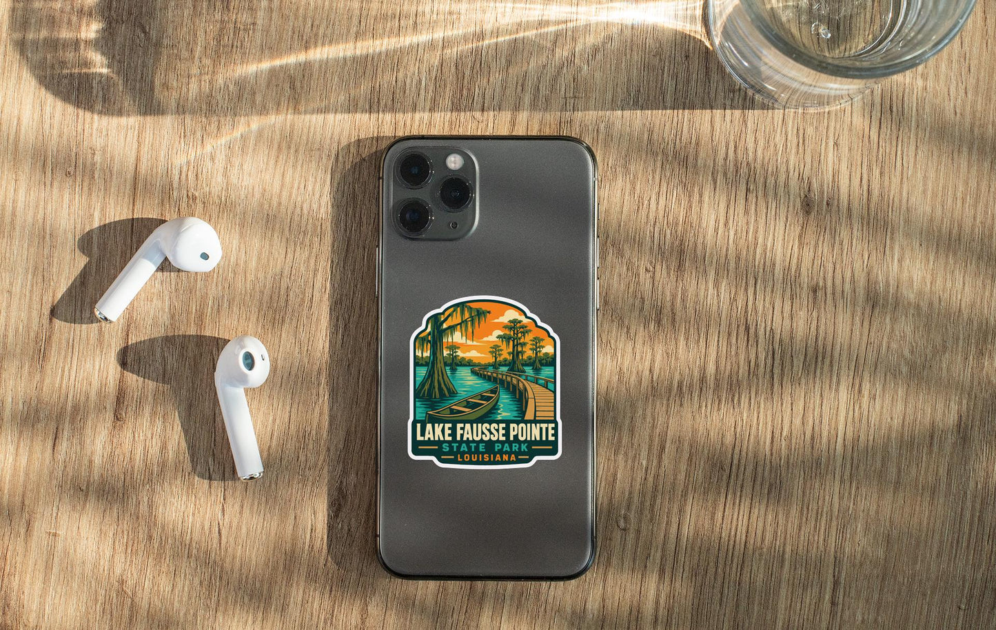 a smartphone with a sticker on the back, placed on a wooden surface alongside a pair of white earbuds and a glass of water.