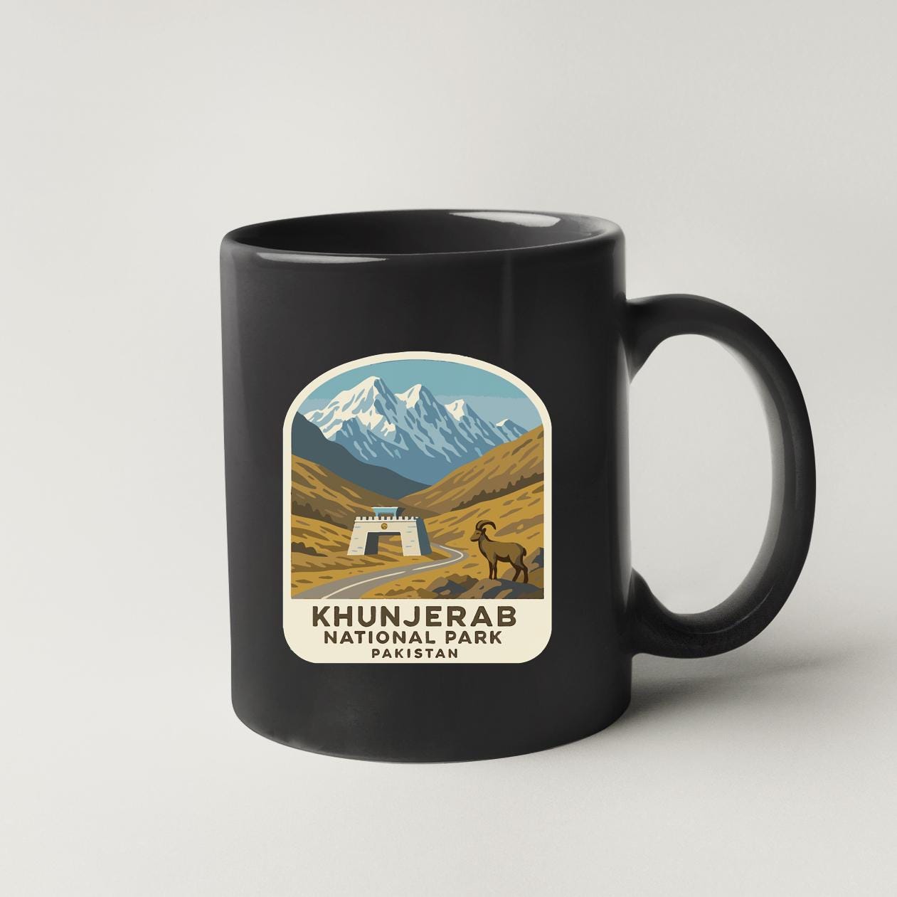 A black coffee mug with a graphic design depicting a mountainous landscape, a goat, and a castle, along with the text "KHUNJERAB NATIONAL PARK PAKISTAN".