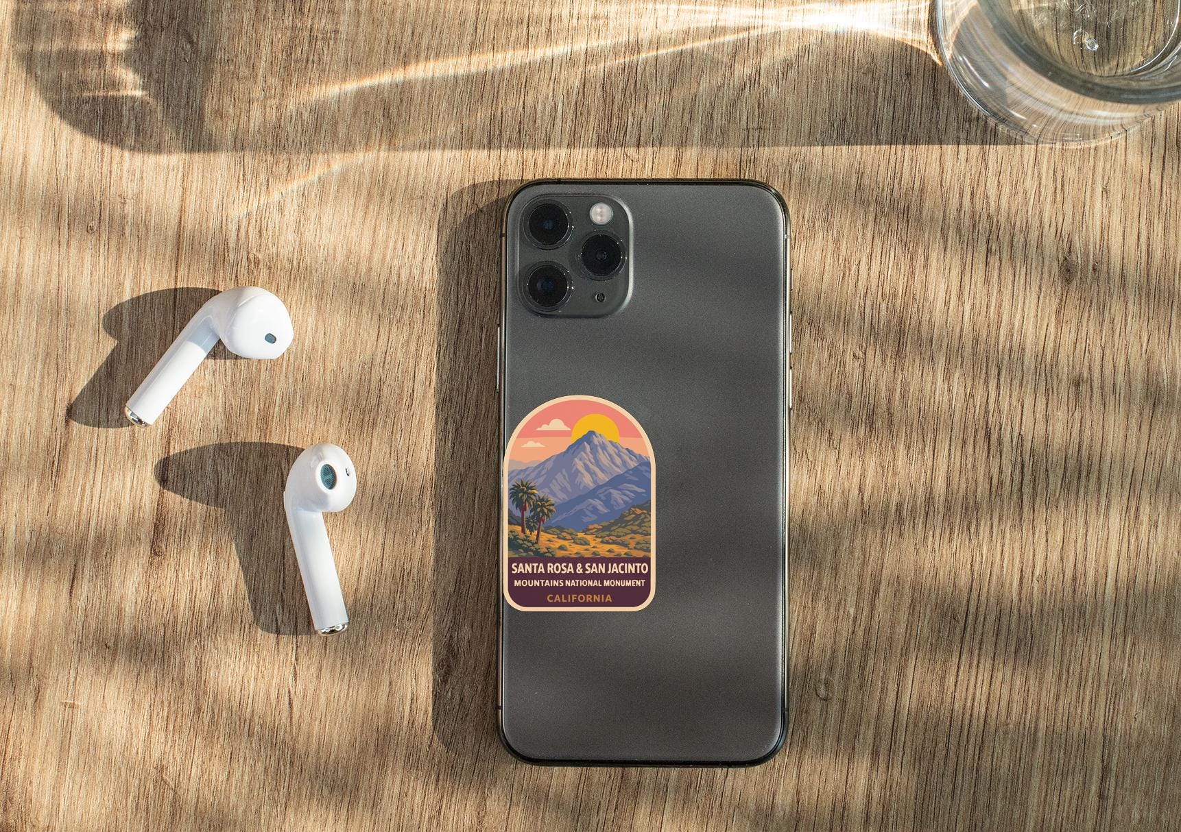 a smartphone with a sticker on the back, placed on a wooden surface alongside a pair of white earbuds and a glass of water.