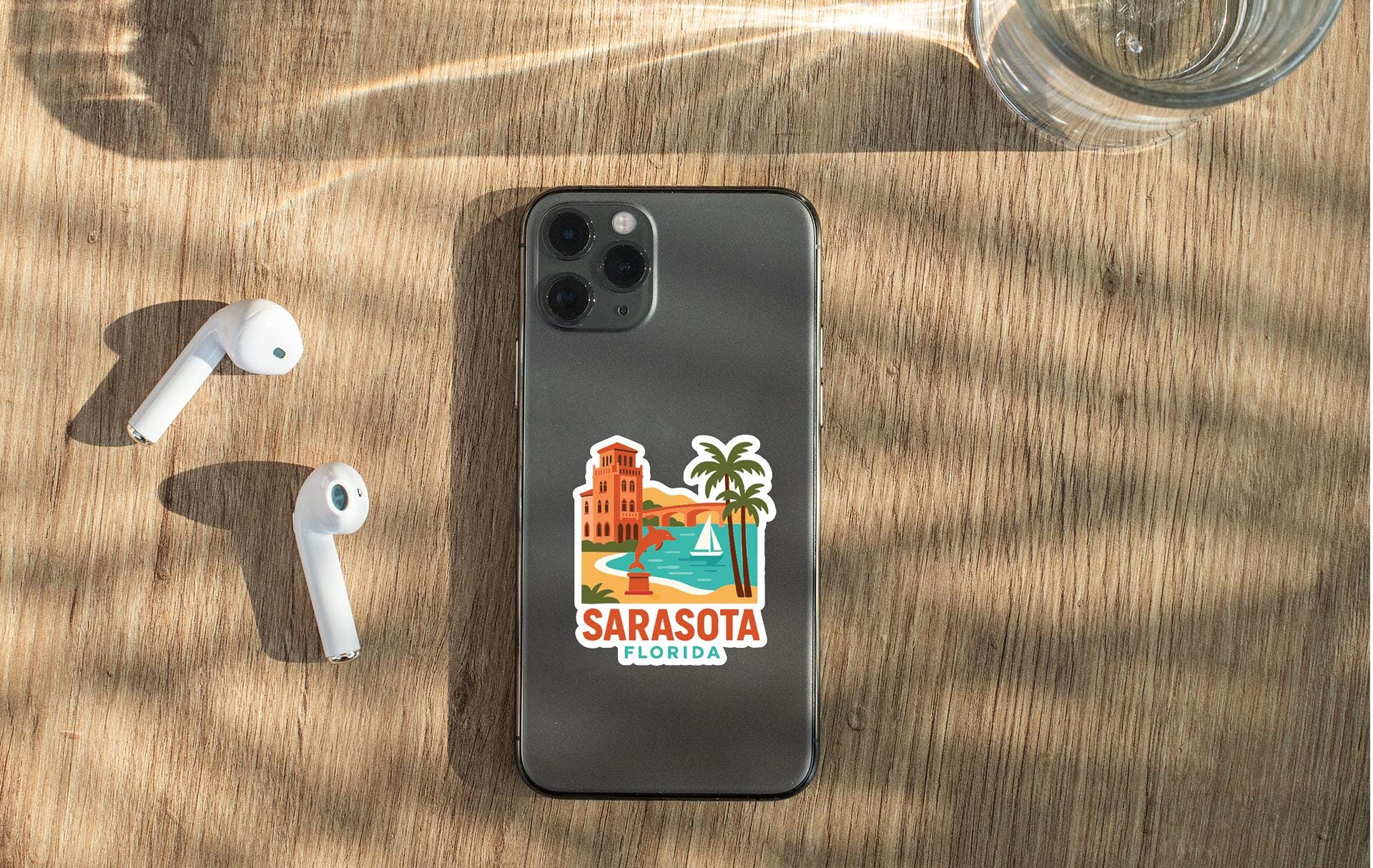 a smartphone with a sticker of a beach and palm trees, along with a pair of white earbuds and a glass of water on a wooden surface.