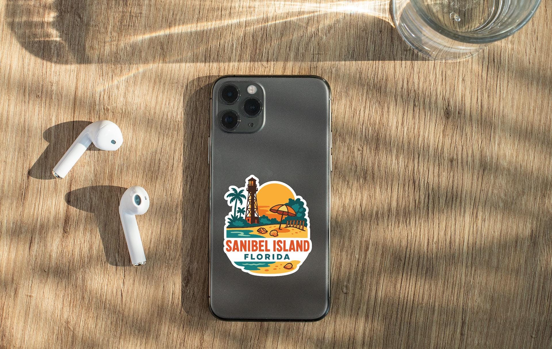 a smartphone with a sticker on the back, placed on a wooden surface alongside a pair of white earbuds and a glass of water.