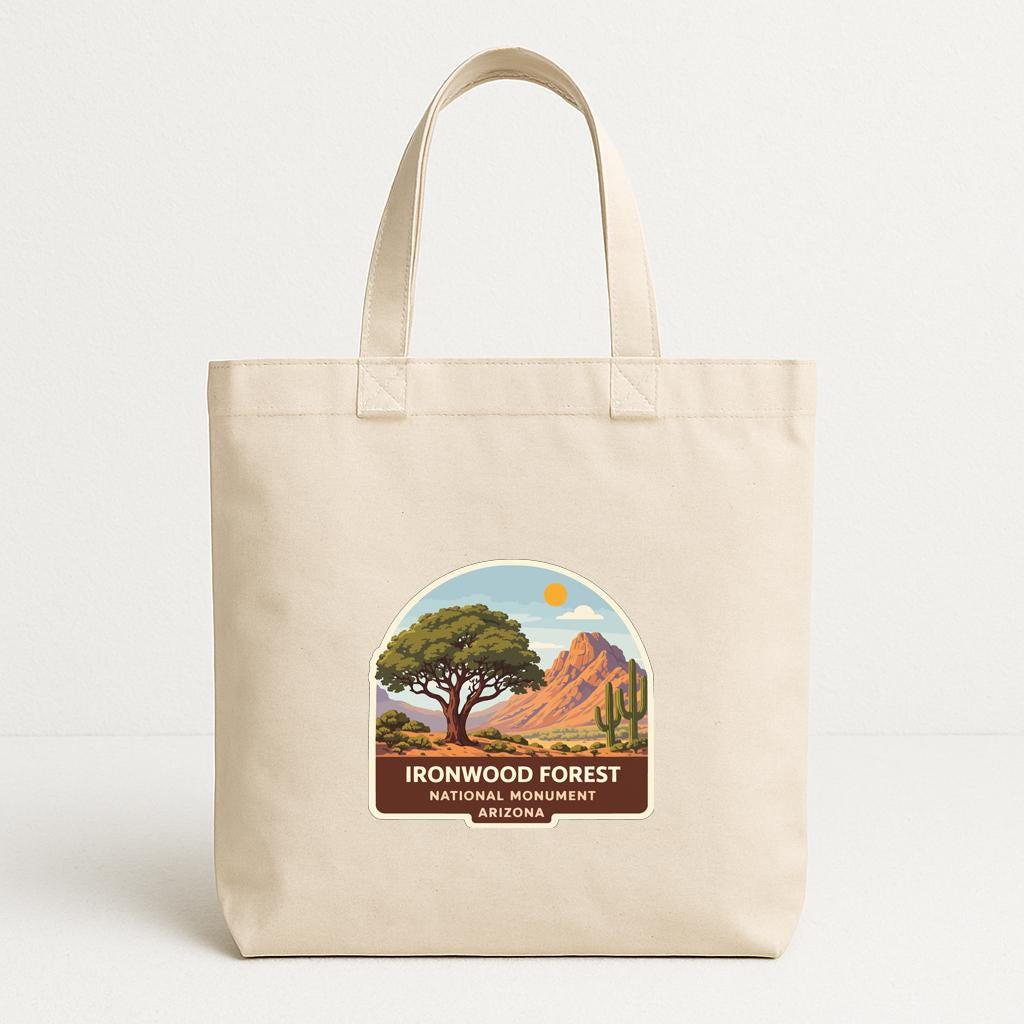 A beige tote bag with a label that says "Ironwood Forest National Monument Arizona" and features a landscape with a tree, mountains, and a desert scene.