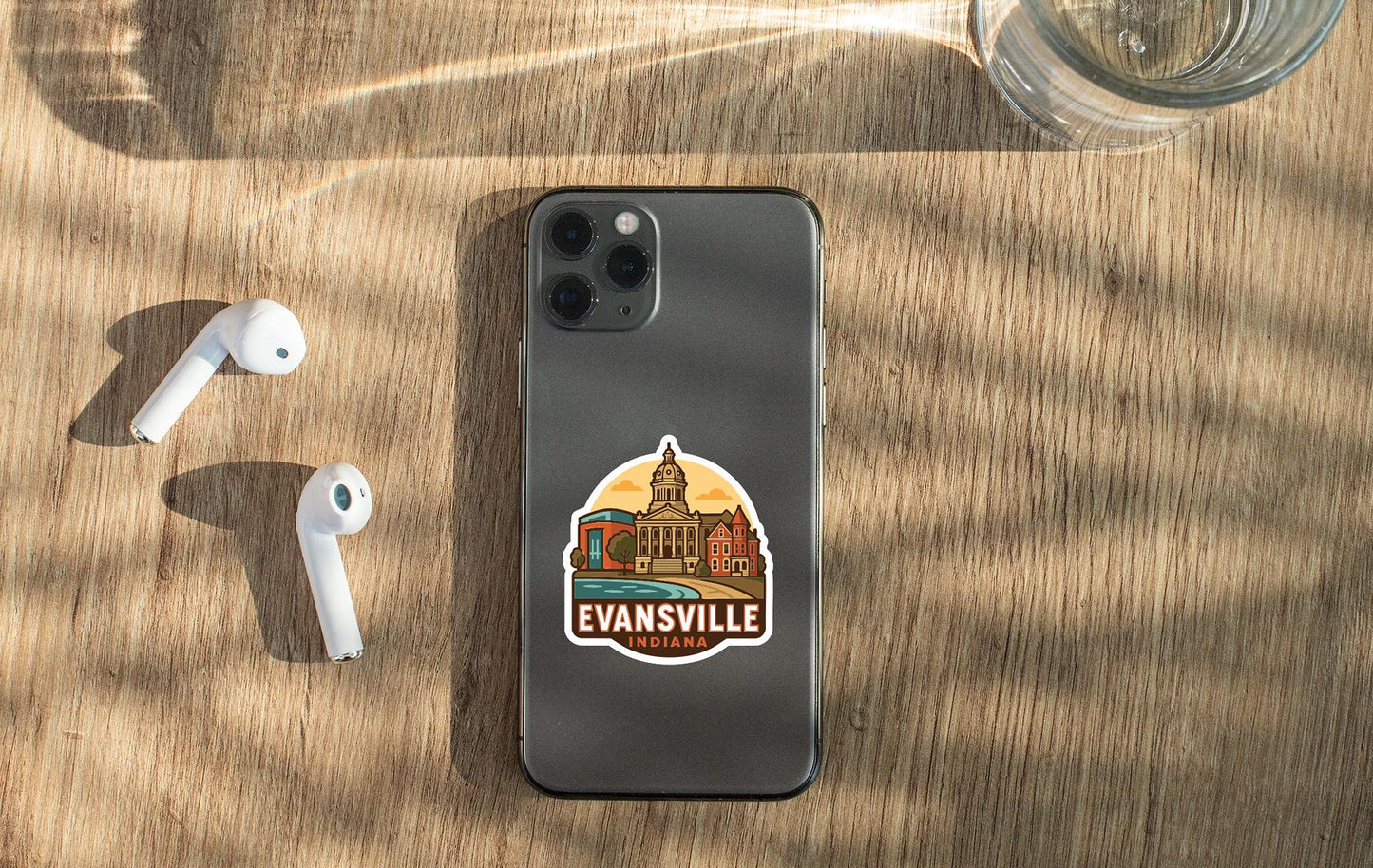 a smartphone with a sticker on the back, along with a pair of white earbuds and a glass of water on a wooden surface.