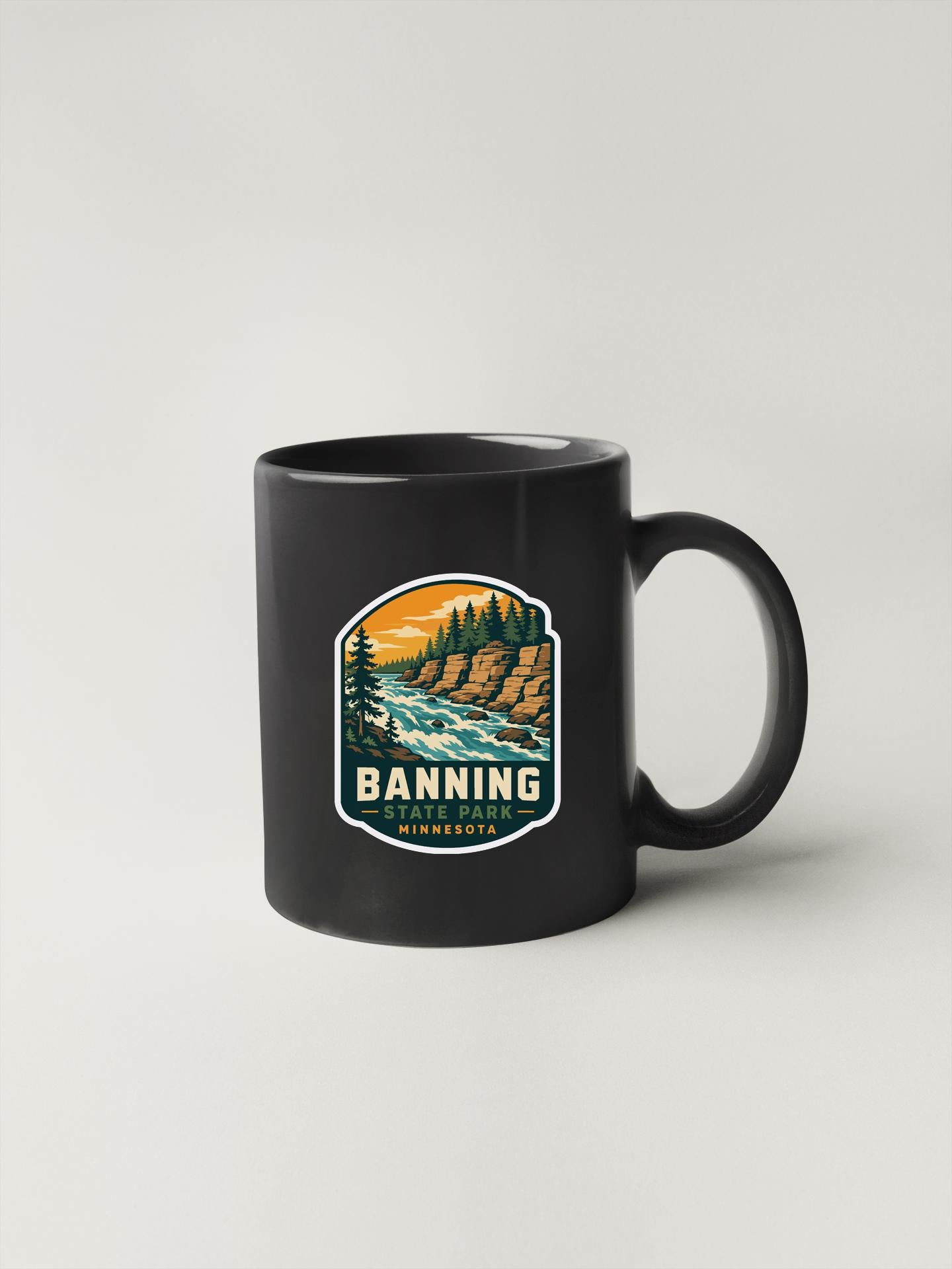 A black coffee mug with a graphic design depicting a scenic landscape with a river, mountains, and pine trees.