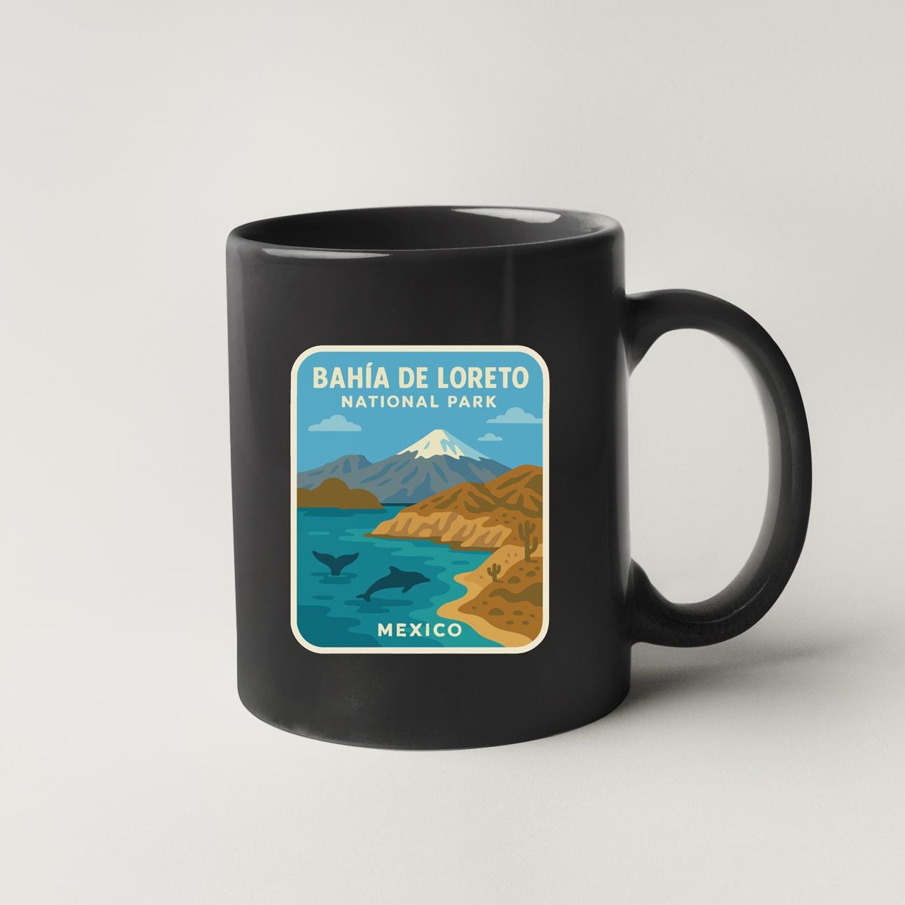 A black coffee mug with a graphic design depicting a scenic landscape with mountains, a lake, and a beach, along with the text "BAHIA DE LORETO NATIONAL PARK MEXICO".