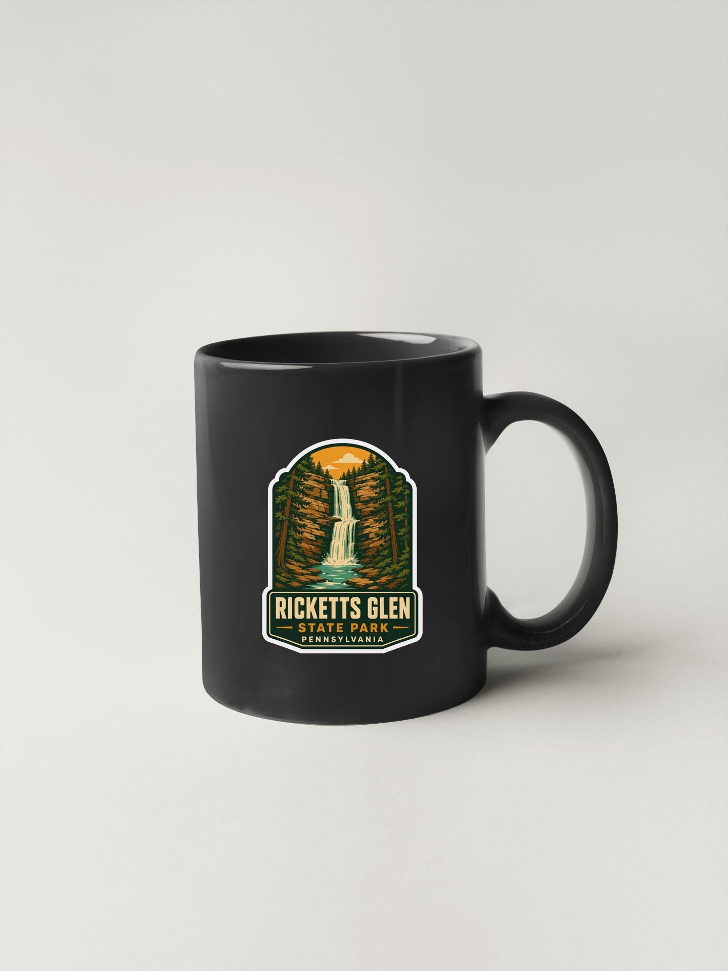 A black coffee mug with a sticker depicting a waterfall and the text "RICKETT'S GLEN STATE PARK PENNSYLVANIA".