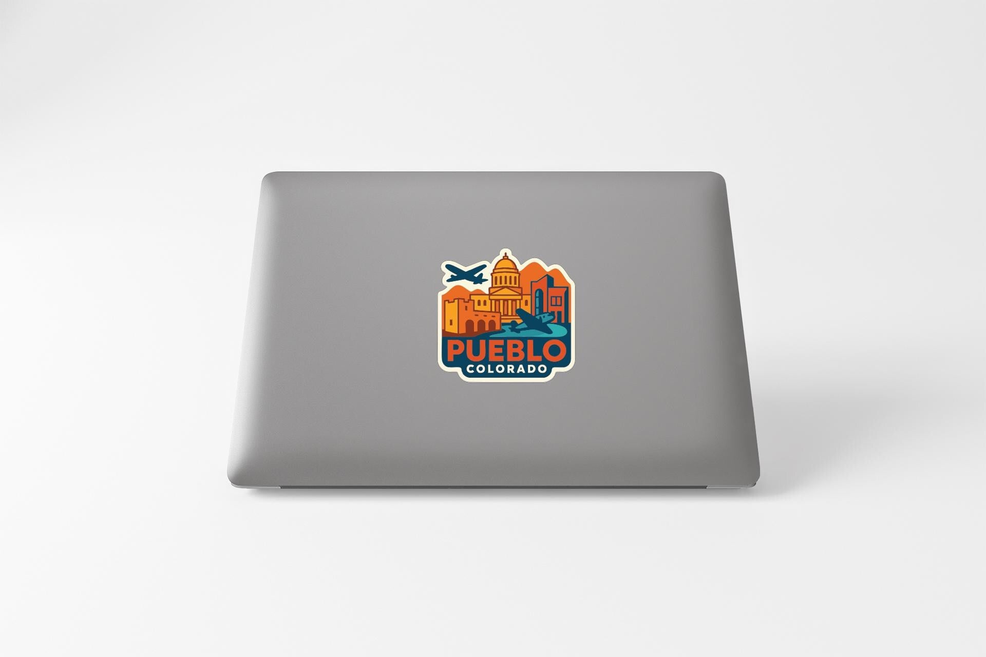 a sticker with a graphic of a city skyline and the text "PUEBLO COLORADO" on a grey laptop.