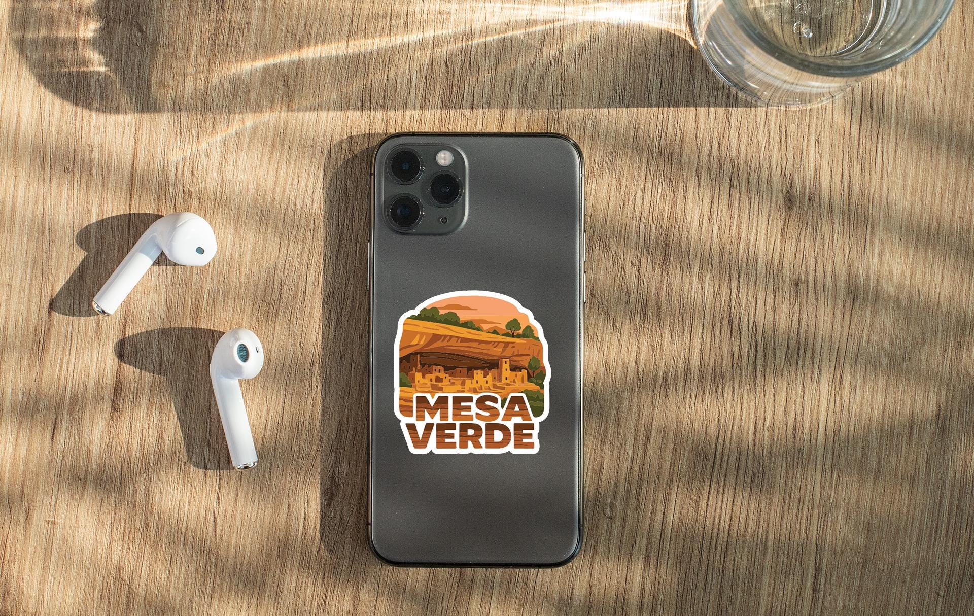 a smartphone with a sticker on the back, placed on a wooden surface alongside a pair of white earbuds and a glass of water.