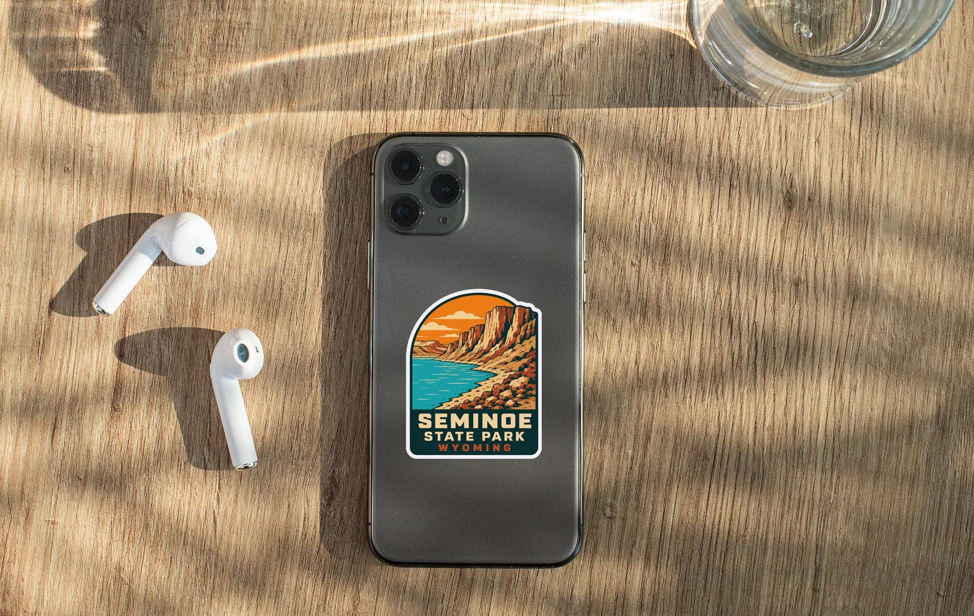 a smartphone with a sticker on the back, placed on a wooden surface alongside a pair of white earbuds and a glass of water.