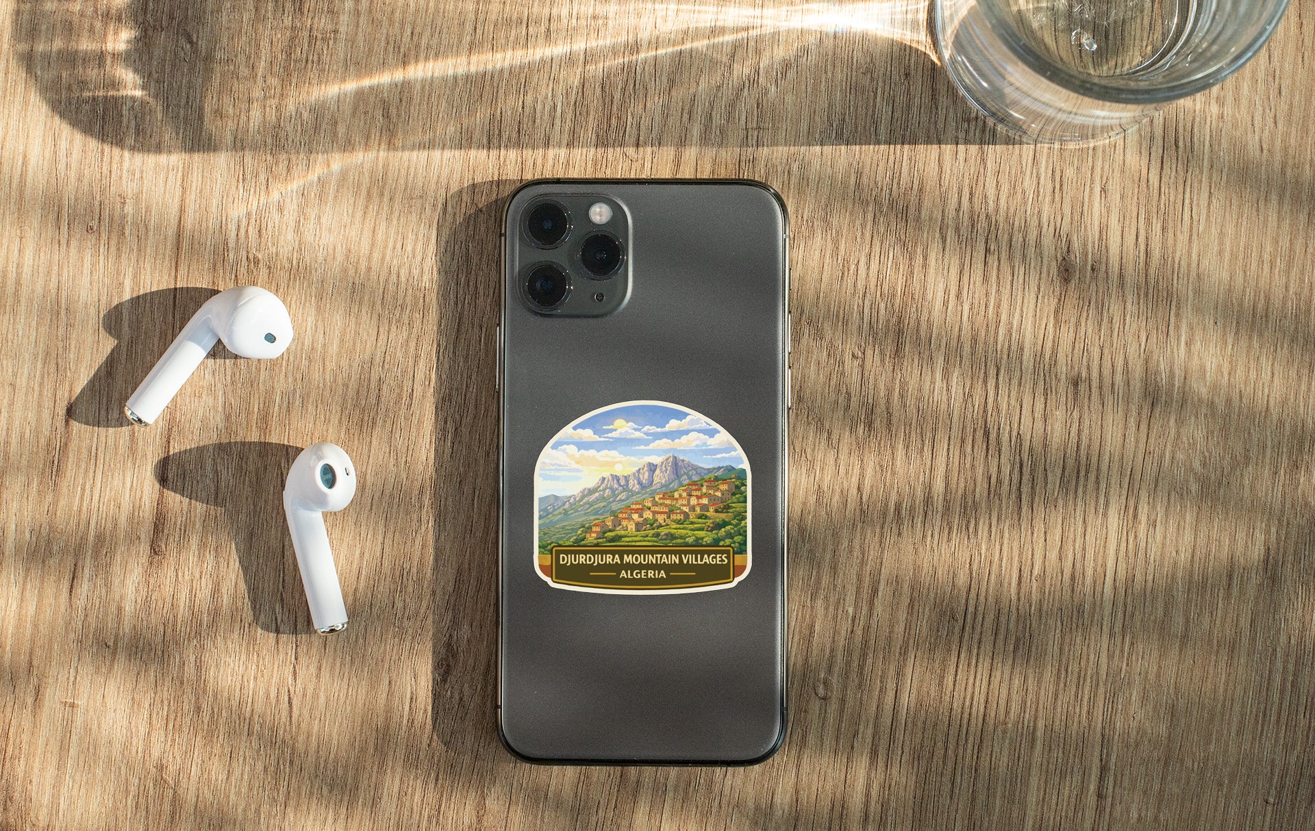 a smartphone with a sticker of a mountain village on its back, along with a pair of white earbuds and a glass of water on a wooden surface.