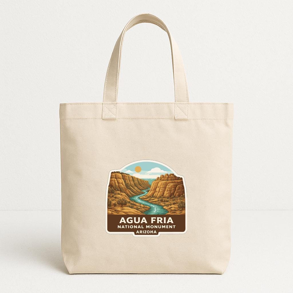 A beige tote bag with a printed label that says "AQUAFRIA NATIONAL MONUMENT ARIZONA" depicting a scenic landscape with a river and mountains.
