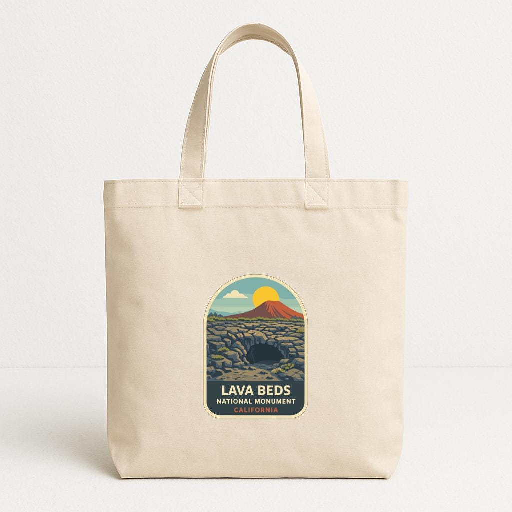 A beige tote bag with a graphic design featuring a mountain landscape and the text "LAVA BEDS NATIONAL MONUMENT CALIFORNIA".