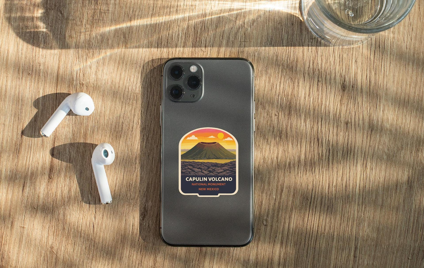 a smartphone with a sticker of a volcano on it, placed on a wooden surface alongside a pair of white earbuds and a glass of water.