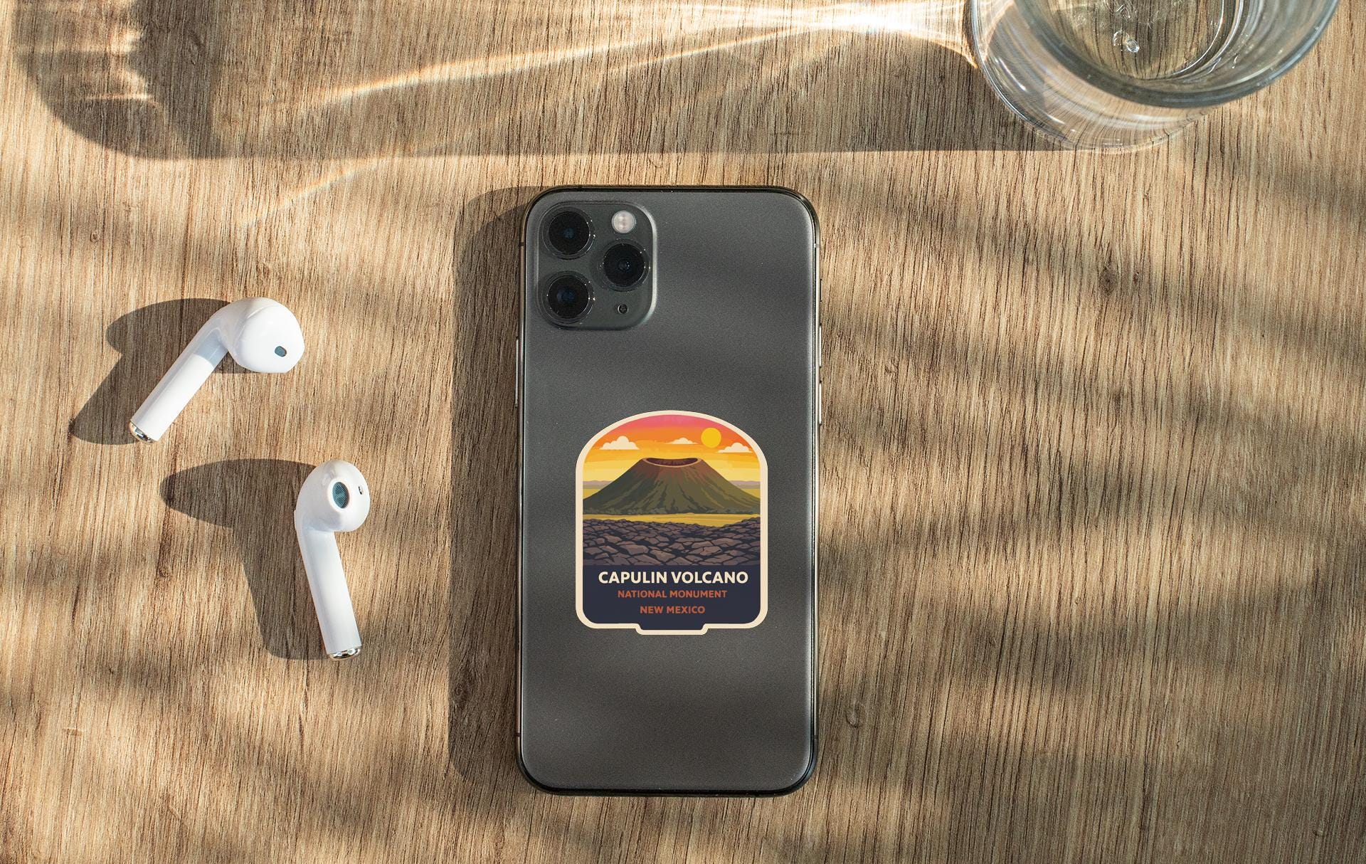 a smartphone with a sticker of a volcano on it, placed on a wooden surface alongside a pair of white earbuds and a glass of water.