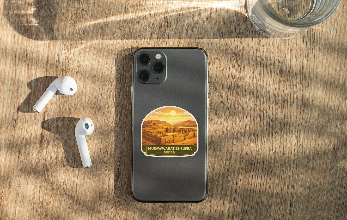 a smartphone with a sticker on the back, a pair of white earbuds, and a glass of water on a wooden surface.