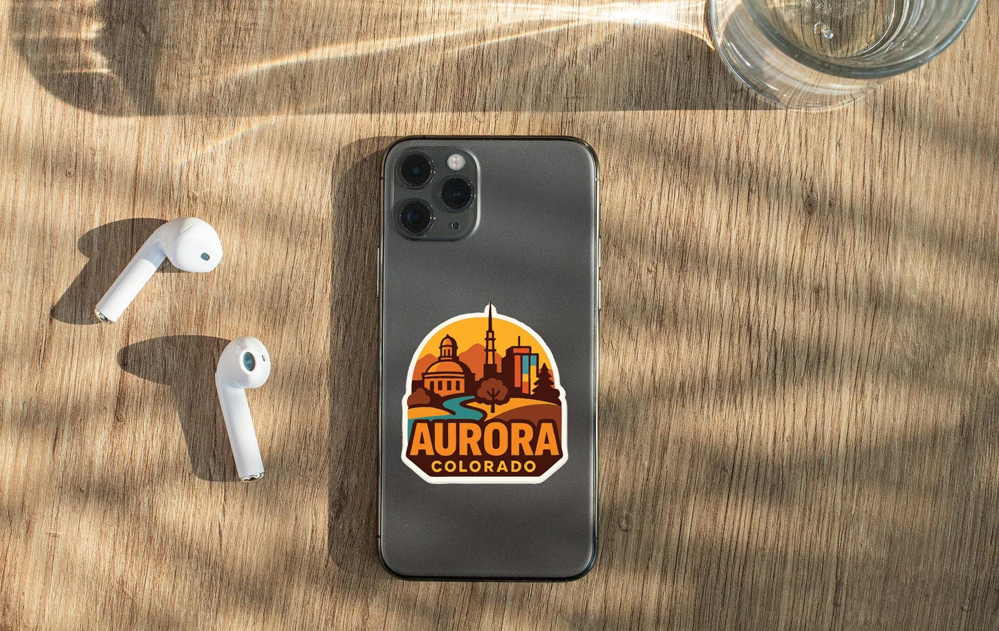 a smartphone with a sticker of Aurora, Colorado, placed on a wooden surface alongside a pair of white earbuds and a glass of water.