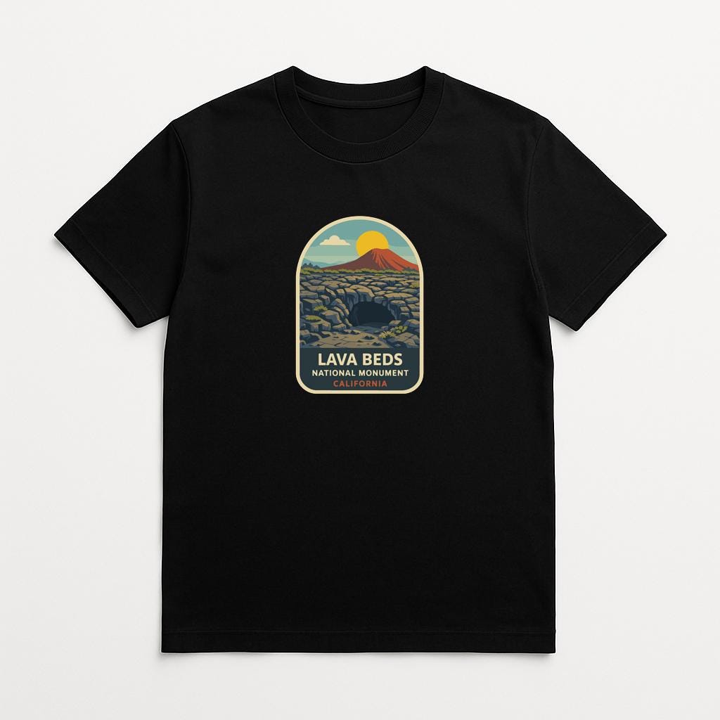 A black t-shirt with a graphic design depicting a landscape with a mountain, a cave, and a sunset. The text below the design reads "LAVA BEDS NATIONAL MONUMENT".