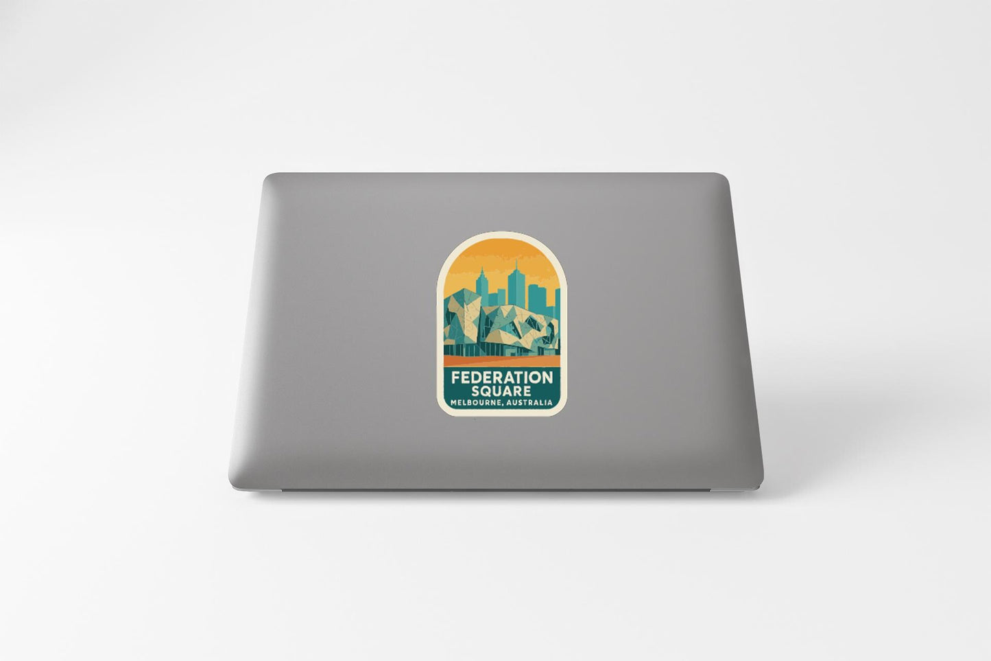 a sticker with a graphic of a cityscape and the text "FEDERATION SQUARE MELBOURNE AUSTRALIA" on a grey laptop.