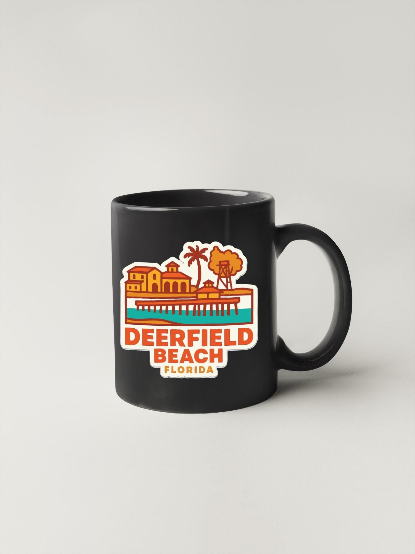 A black coffee mug with a colorful sticker depicting Deerfield Beach, Florida.