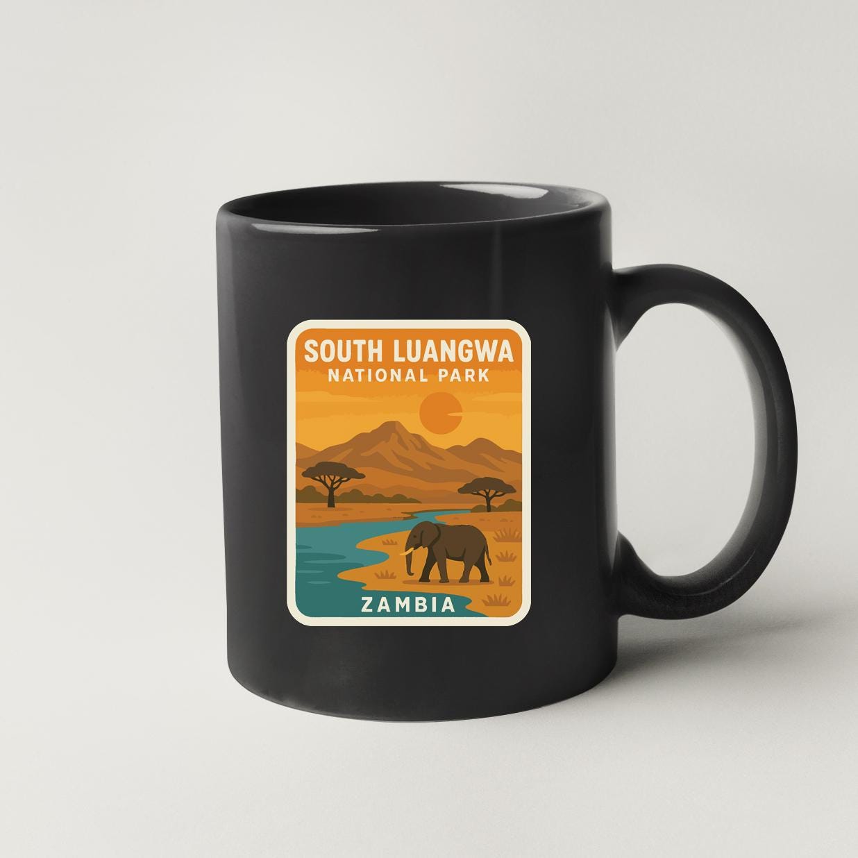 A black coffee mug with a graphic design depicting a landscape with a river, mountains, and an elephant, along with the text "SOUTH LUANGWA NATIONAL PARK ZAMBIA".