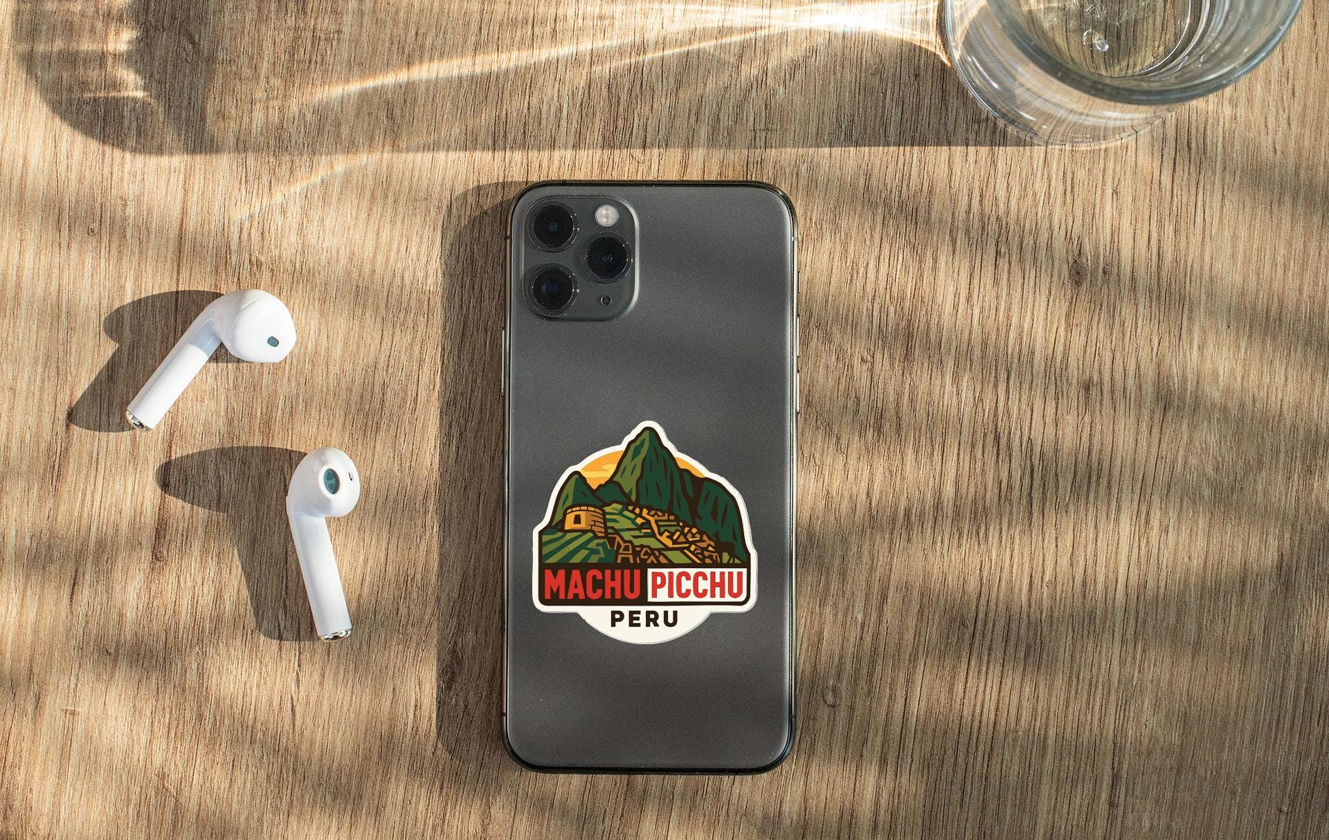a smartphone with a sticker on the back, placed on a wooden surface alongside a pair of white earbuds and a glass of water.