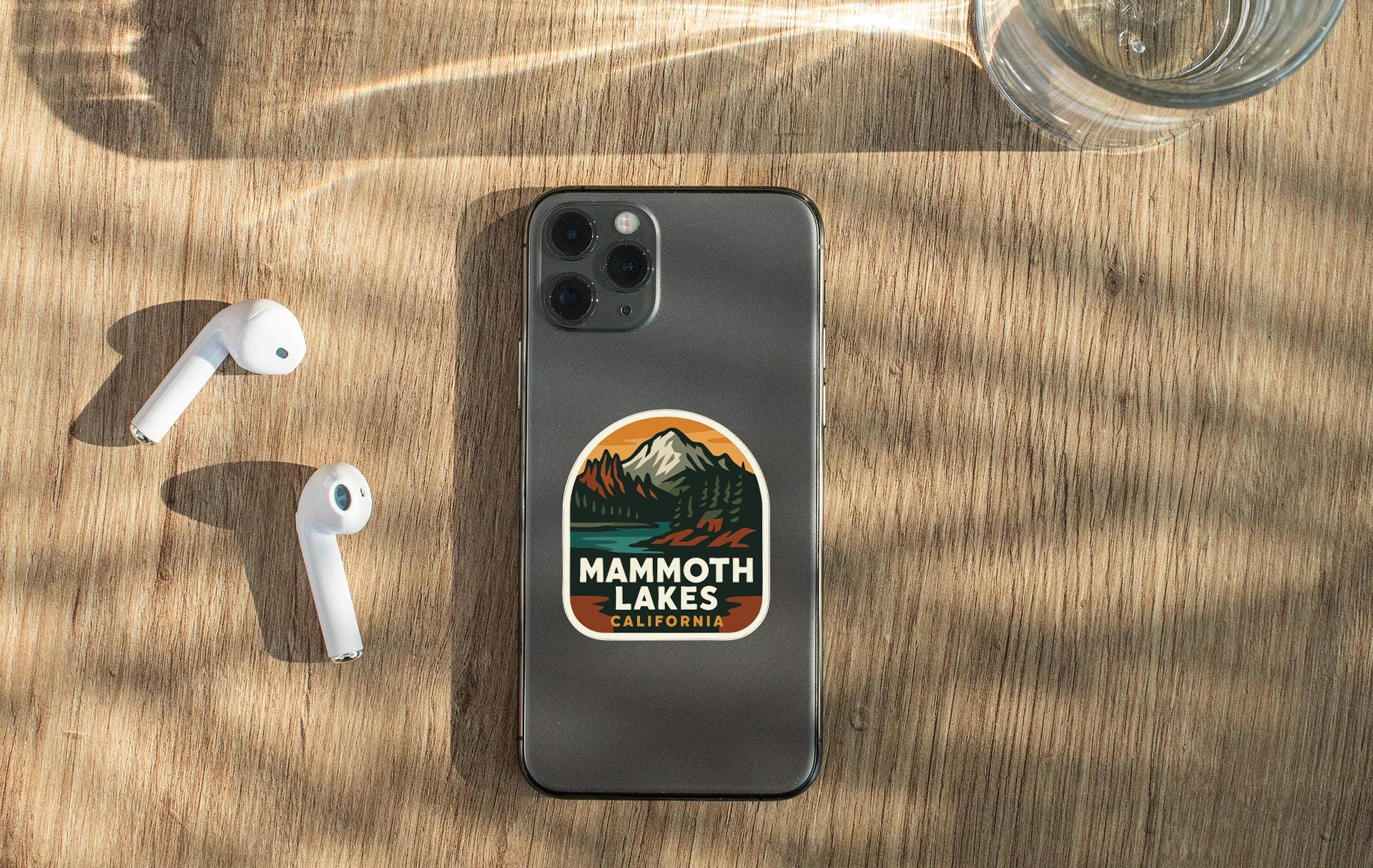 a smartphone with a sticker on the back, placed on a wooden surface alongside a pair of white earbuds and a glass of water.